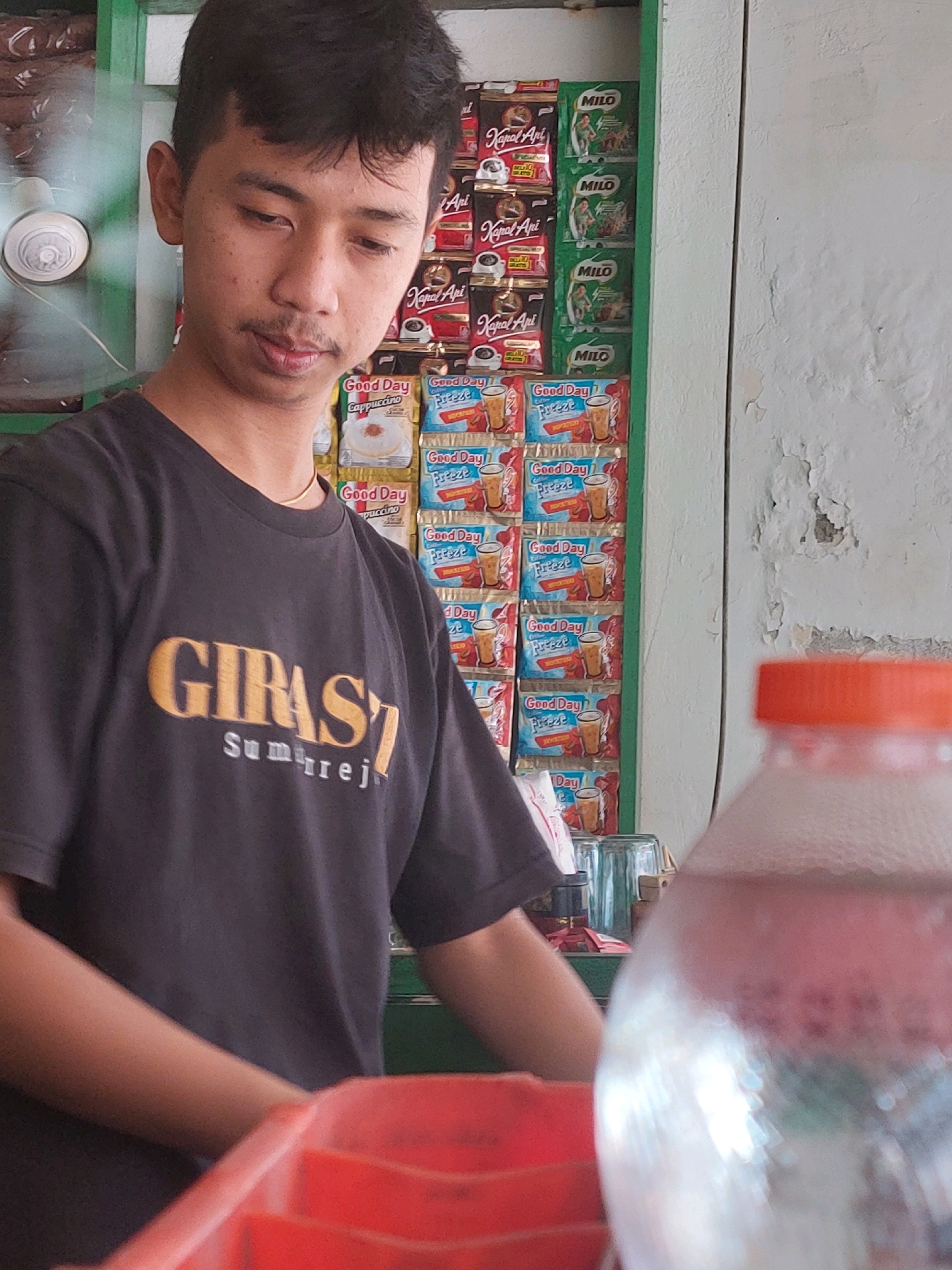 A young man with dark hair and a thin mustache is standing in what appears to be a small store or kiosk in Sumuragung, Indonesia. He is wearing a dark t-shirt with a yellow and white logo that reads "GIRAS7 SUMURREJO". Behind him, shelves are stocked with various packaged food and beverage items, including "Good Day" coffee sachets and "Milo" drink packets. A white oscillating fan is visible on the far left. In the foreground, a portion of a clear plastic bottle with an orange cap is in focus, and a red plastic container is partially visible at the bottom. The wall behind the shelves is painted white and shows signs of peeling paint. The overall lighting suggests an indoor, possibly shaded environment.