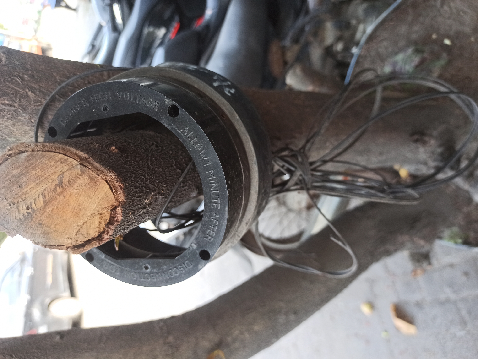 A close-up shot focuses on a segment of a tree trunk in a city environment, possibly in Medan, Indonesia, given the context. A black, circular object, likely a component of electrical equipment due to the inscription "DANGER HIGH VOLTAGE" and "ALLOW 1 MINUTE AFTER DISCONNECT FOR", is secured around the trunk. This object appears to be made of plastic and has several circular holes. Black wires are loosely attached to the object and hang down, entwined with the tree's branches. In the blurred background, a dark-colored car is visible, as are parts of motorcycles and what appears to be a paved surface with scattered debris, such as leaves and small yellow objects. The overall scene suggests an outdoor urban setting where electrical infrastructure might be integrated with natural elements.