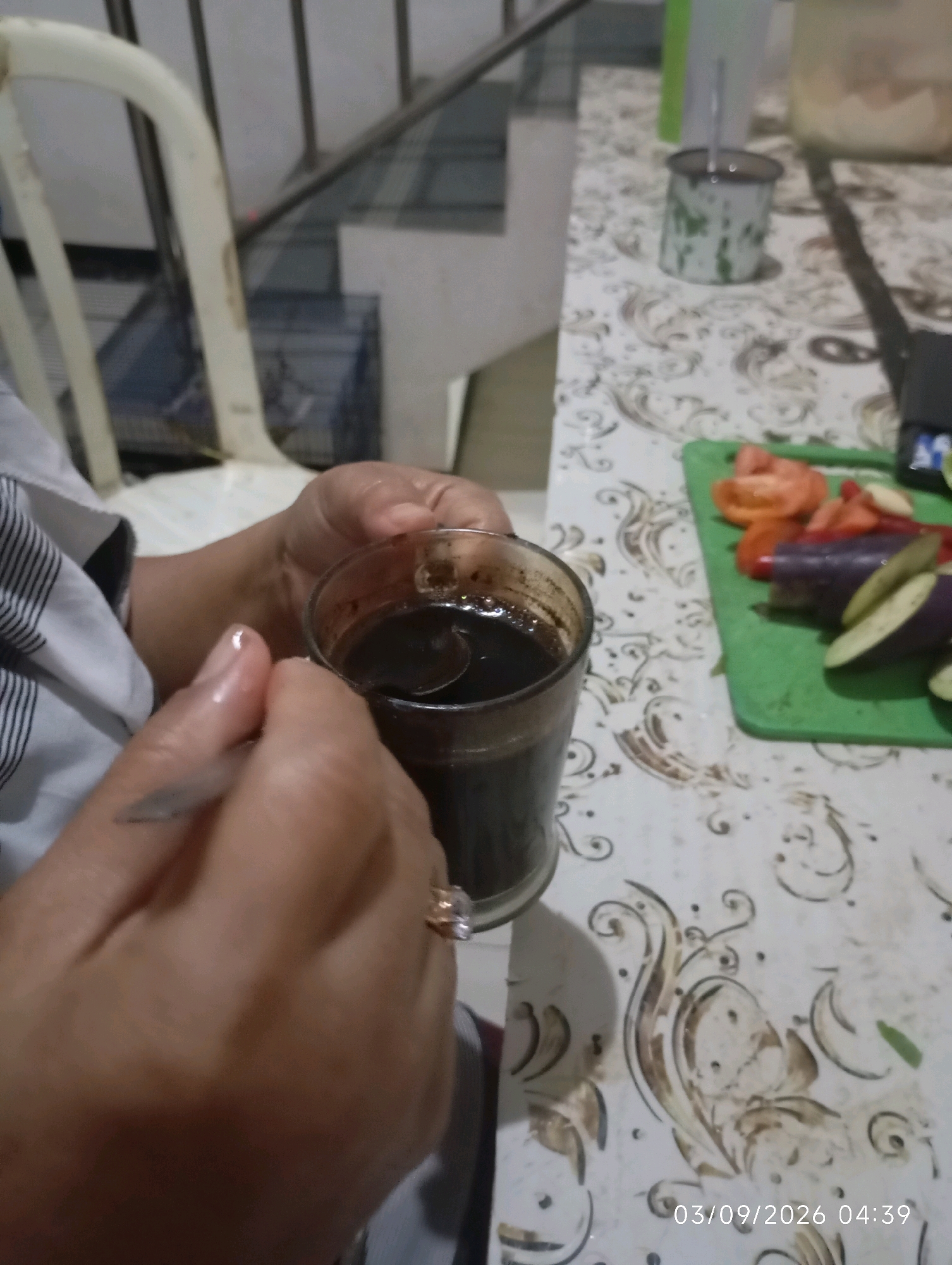 The image shows a close-up view of a person's hands holding a glass of dark liquid, possibly coffee or tea, with a spoon inside. The person is wearing a striped shirt and a ring on their left hand. In the background, a cutting board with sliced tomatoes and eggplant is visible, along with a metal cup and a white chair. The scene appears to be indoors, possibly a kitchen or dining area, with a patterned tablecloth on the table. The timestamp "03/09/2026 04:39" is present in the lower right corner of the image. The location is specified as Klecorejo, Indonesia.