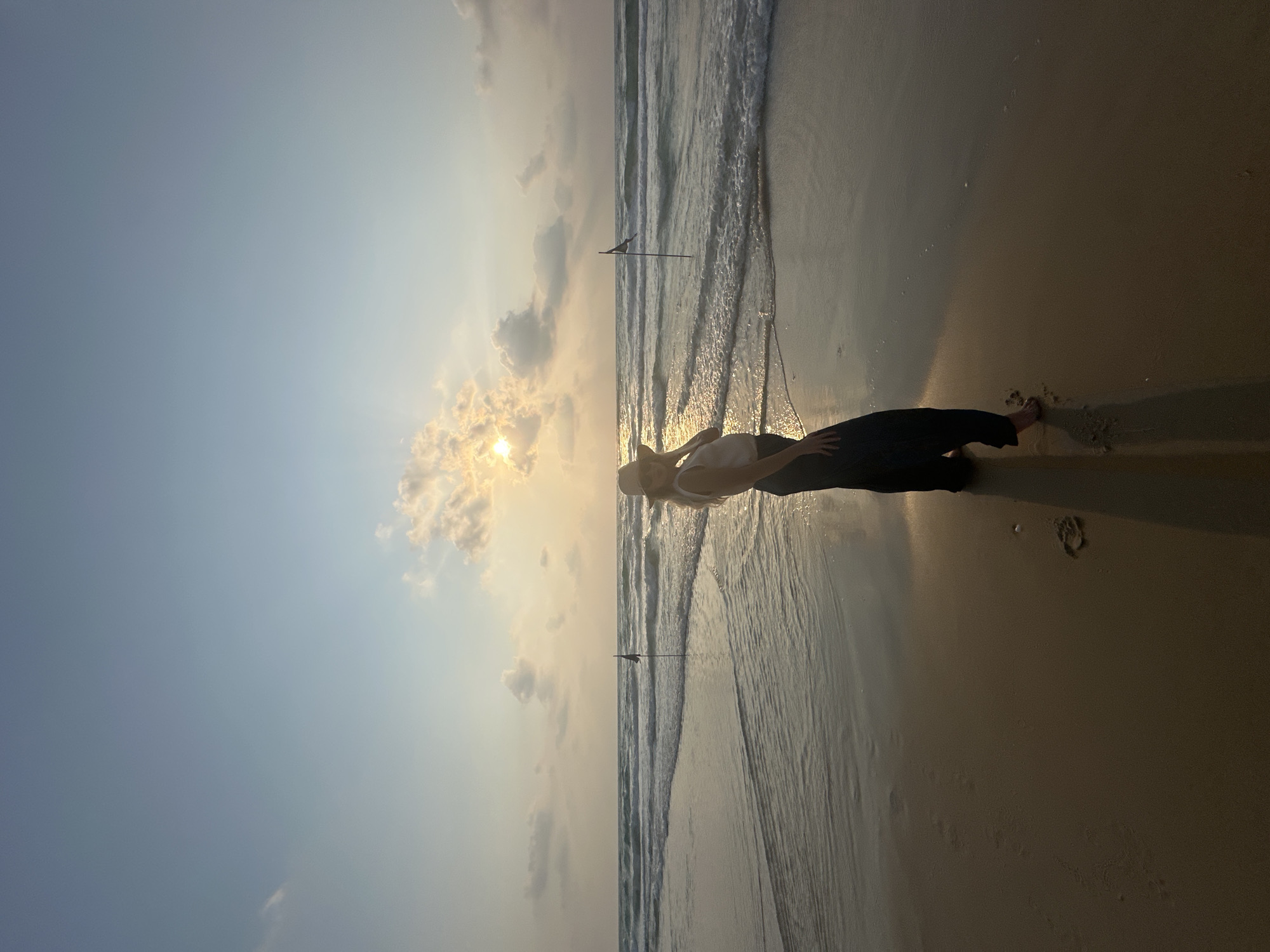 A full-body, low-angle, vertical shot captures a woman standing on the shore of Bat Yam, Israel, during what appears to be sunset. The woman, facing to her right and slightly away from the camera, is wearing a white tank top, dark pants, and a wide-brimmed hat. Her blonde hair is visible beneath the hat. She has one hand on her hip and the other resting on her hat. Her shadow stretches out before her on the wet sand.
The background features the calm ocean with gentle waves rolling onto the beach. The sun is partially obscured by clouds in the upper center of the frame, casting a warm, golden light across the sky and water. Two dark flags on poles are visible in the distance, one closer to the shore and the other further out in the water. The sand is wet and reflective, with ripples from the receding waves. The overall atmosphere is serene and tranquil.