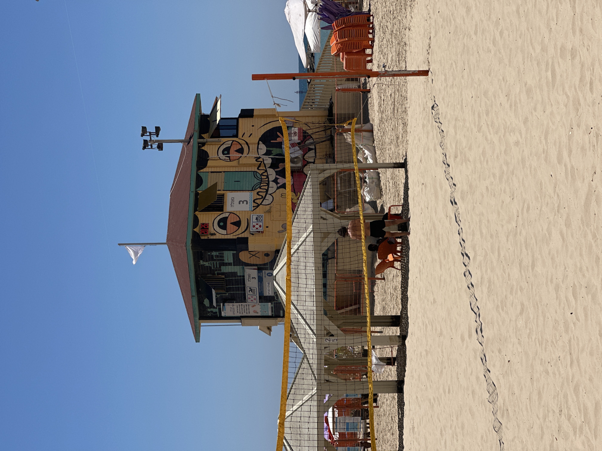 The image captures a vibrant beach scene under a clear, bright blue sky. In the foreground and stretching towards the background is a sandy beach, marked with numerous footprints.
The central focus is a colorful, two-story wooden beach shack or lifeguard station, adorned with large, cartoon-like faces and graffiti-style artwork. A white flag flies from a pole on its roof. Attached to the shack, and extending into the sand, is a yellow and black volleyball net, dividing the sandy area. Orange chairs are stacked near the shack and scattered beside the net, suggesting a relaxation or waiting area.
On the right side of the frame, partially obscured by the net and chairs, is a person wearing dark shorts, seemingly adjusting something near the net. Another person's legs and feet are visible further to the right. Beyond the shack, in the distance, a calm expanse of blue sea is visible under a white and purple beach umbrella.
The bright sunlight indicates it is daytime, likely midday or early afternoon, with sharp shadows visible on the sand and objects. The clear sky and absence of clouds suggest fair weather.
Visible text on the wooden shack includes:
* A large number "3" with Hebrew text "מאלכד" (Malched/Ma'alched) above it.
* A white sign with Hebrew text "שעות פעילות" (Operating Hours) followed by the time "07:15-17:45".
* Below this, "טלפון" (Telephone) and a number "03-411-5941".
* Other smaller signs with symbols (e.g., life preserver, no smoking, no dogs) and illegible Hebrew text are also present on the shack.