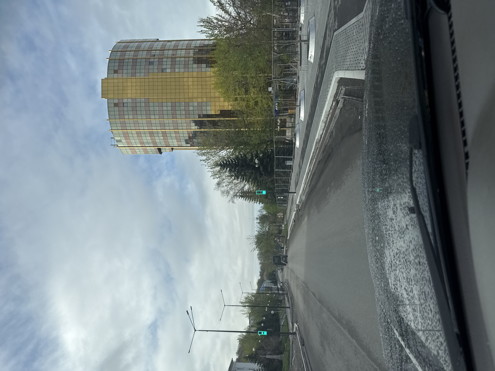 The image depicts a street scene in Petropavlovsk-Kamchatsky, Russia. A multi-story, curved building with a gold-colored facade is visible on the left side of the frame. The street stretches away from the viewer and is lined with trees and streetlights. Traffic lights are also visible on the street. The photo was taken from inside a vehicle, with part of the dashboard visible in the foreground. The sky is cloudy.
