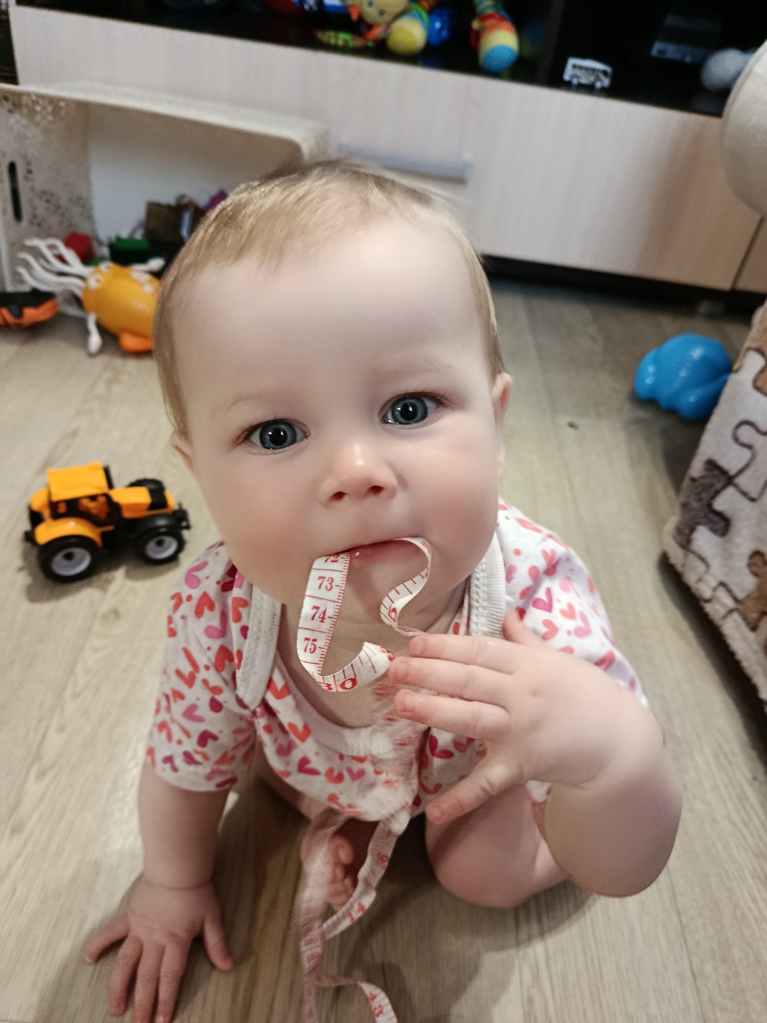A fair-skinned baby with blue eyes is sitting on a light wood-patterned floor. The baby is wearing a white onesie with red and pink heart patterns. The baby has a white tape measure in its mouth, with the numbers 73, 74, and 75 visible. The baby's right hand is near its mouth, and a portion of the tape measure is draped around its torso. Various toys are visible in the background, including an orange and black toy truck to the left, and other toys near a white shelf and a dark brown cabinet in the upper part of the frame. A blue toy is visible to the right, near a patterned blanket or cover. The location is Unknown, Unknown.