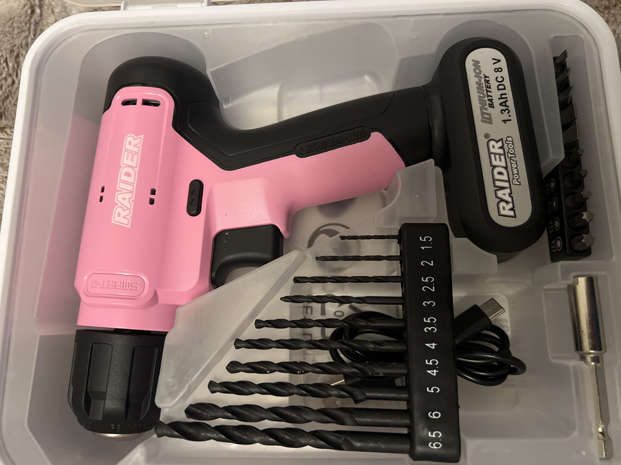 The image shows a pink and black cordless drill set inside a white plastic case. The drill, labeled "RAIDER SMART-D," features a black handle and a battery pack. Next to the drill are various drill bits, a USB charging cable, and an adapter for screwdriver bits. The set appears new, neatly organized within the container. The location in Sofia, Bulgaria, is not mentioned or implied in the image.