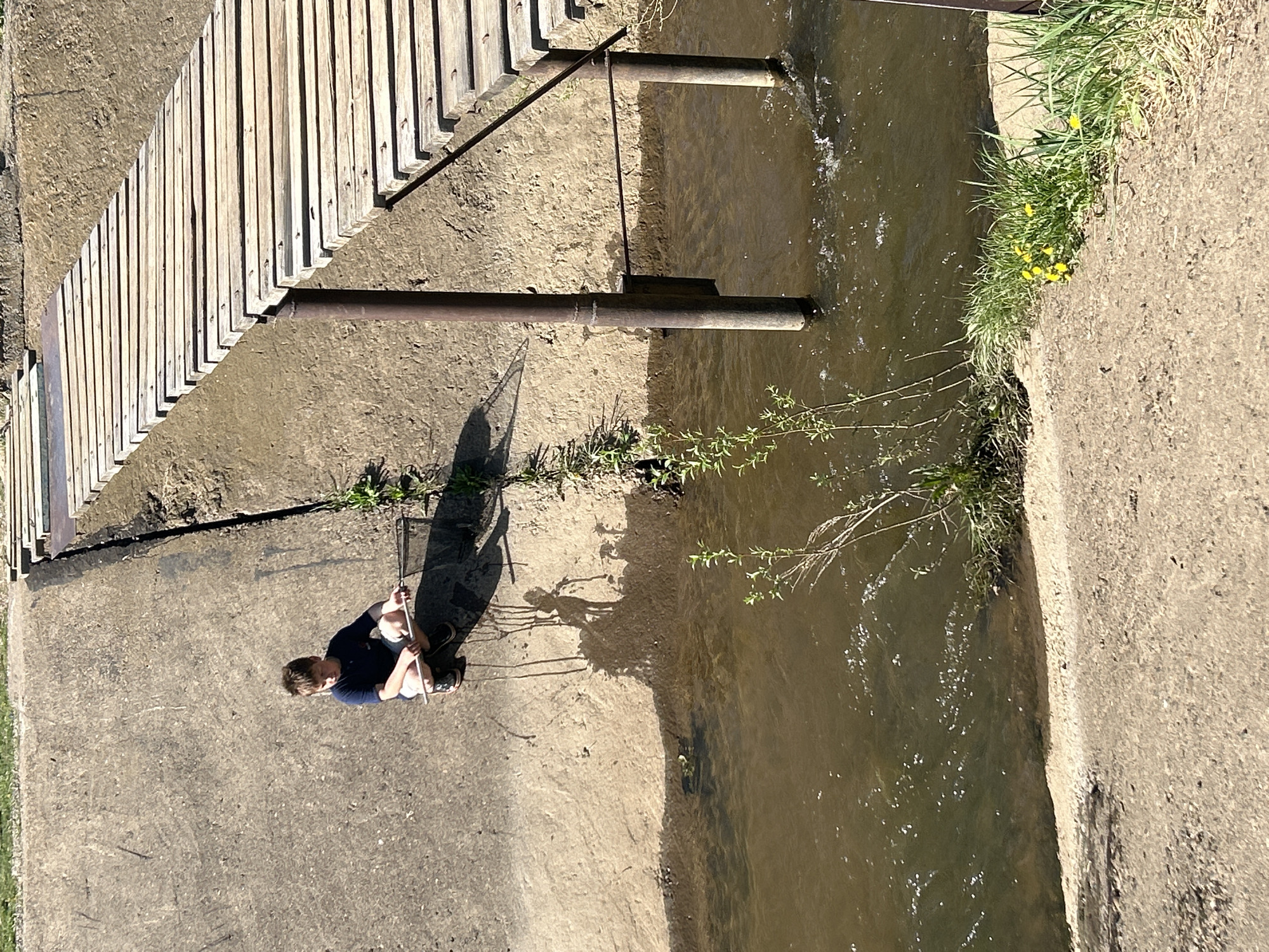 A medium-angle shot shows a boy seated on a concrete path beside a canal, possibly located in Chaikovsky, Russia. The path is adjacent to the water's edge, where some vegetation grows. A wooden structure and metal pipes cross over the canal. The boy is holding a stick.