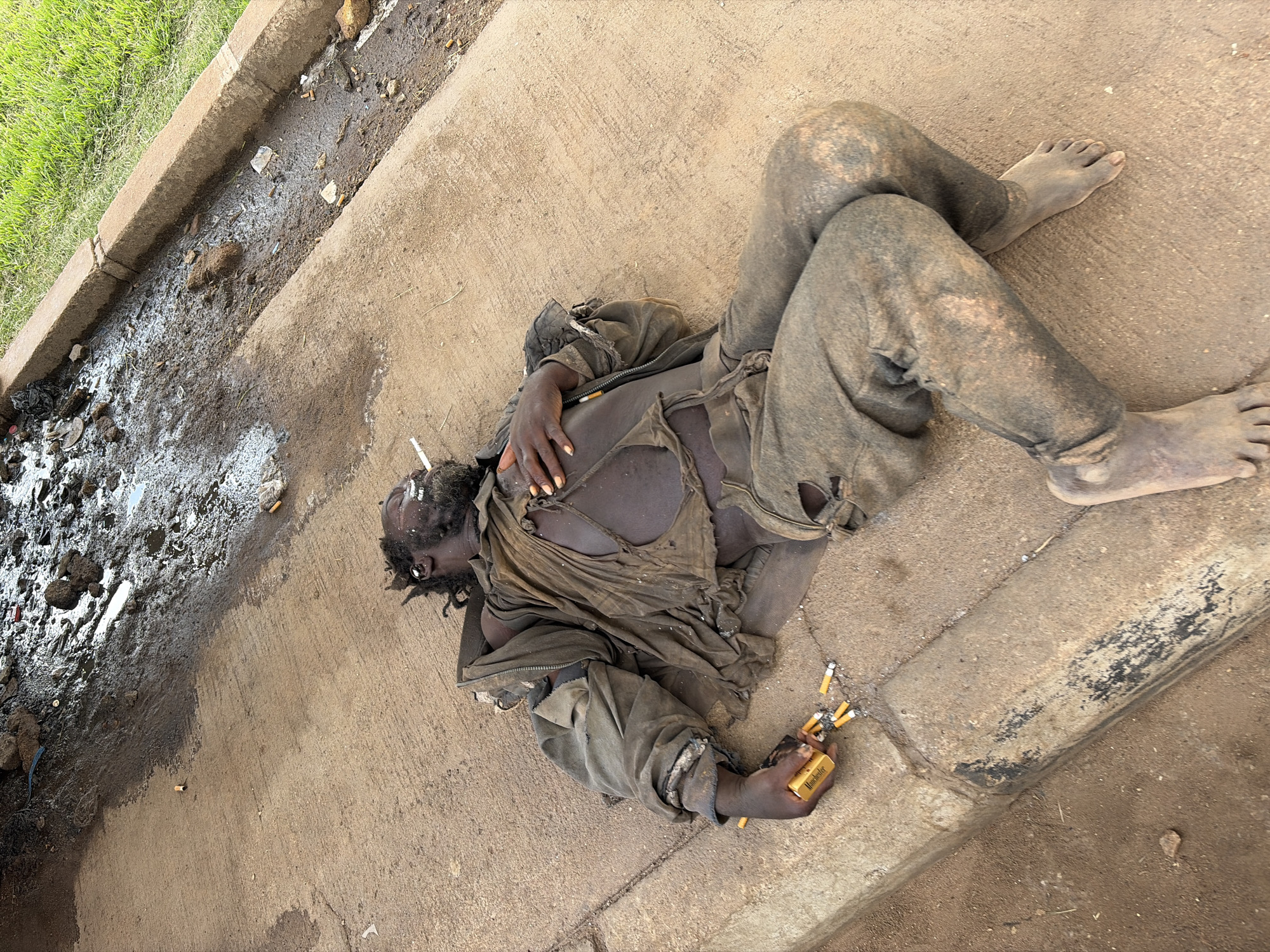 An individual lies on their back on a concrete surface, appearing to be asleep or unconscious. They are dressed in torn, dirty, dark-colored clothing, with rips visible in their shirt and pants. Their skin is dark, and their body is also covered in dirt and grime. The individual's face is partially obscured by their hair, which appears dark and possibly matted.
In their right hand, the individual is holding a pack of cigarettes, with several loose cigarettes also scattered around their hand and on the ground. A lit cigarette is visible in their mouth.
The concrete surface appears to be a sidewalk or a paved area next to a grassy verge and a drainage ditch. The ditch is filled with murky water, debris, and numerous cigarette butts. The overall scene suggests a gritty, possibly urban or semi-urban environment. The lighting suggests it is daytime, with no specific indication of the weather. The individual's posture and the presence of cigarettes could imply a state of exhaustion, intoxication, or homelessness.
The cigarette pack has visible text on it, which appears to be "Manchester".