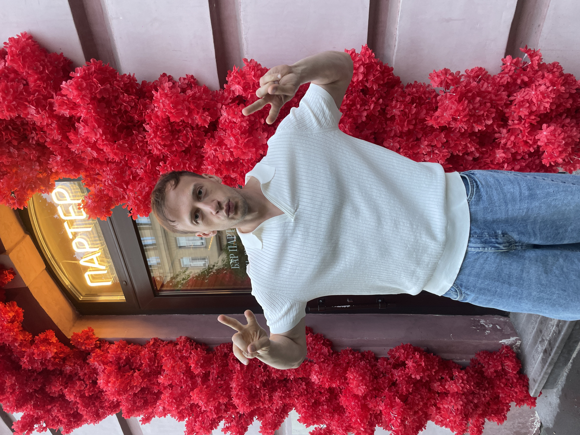A man stands in front of a building decorated with vibrant red floral arrangements, with a neon sign in Russian that reads "ПАРТЕР" (PARTER) above a doorway. He is wearing a white polo shirt and light blue jeans. He is making a peace sign with both hands. The building appears to be a bar or establishment, possibly located in Saint Petersburg, Russia, given the Cyrillic signage. The facade of the building is a light pink color. The man is looking towards the camera with his lips pursed. The red floral decorations are densely packed and appear to be artificial.