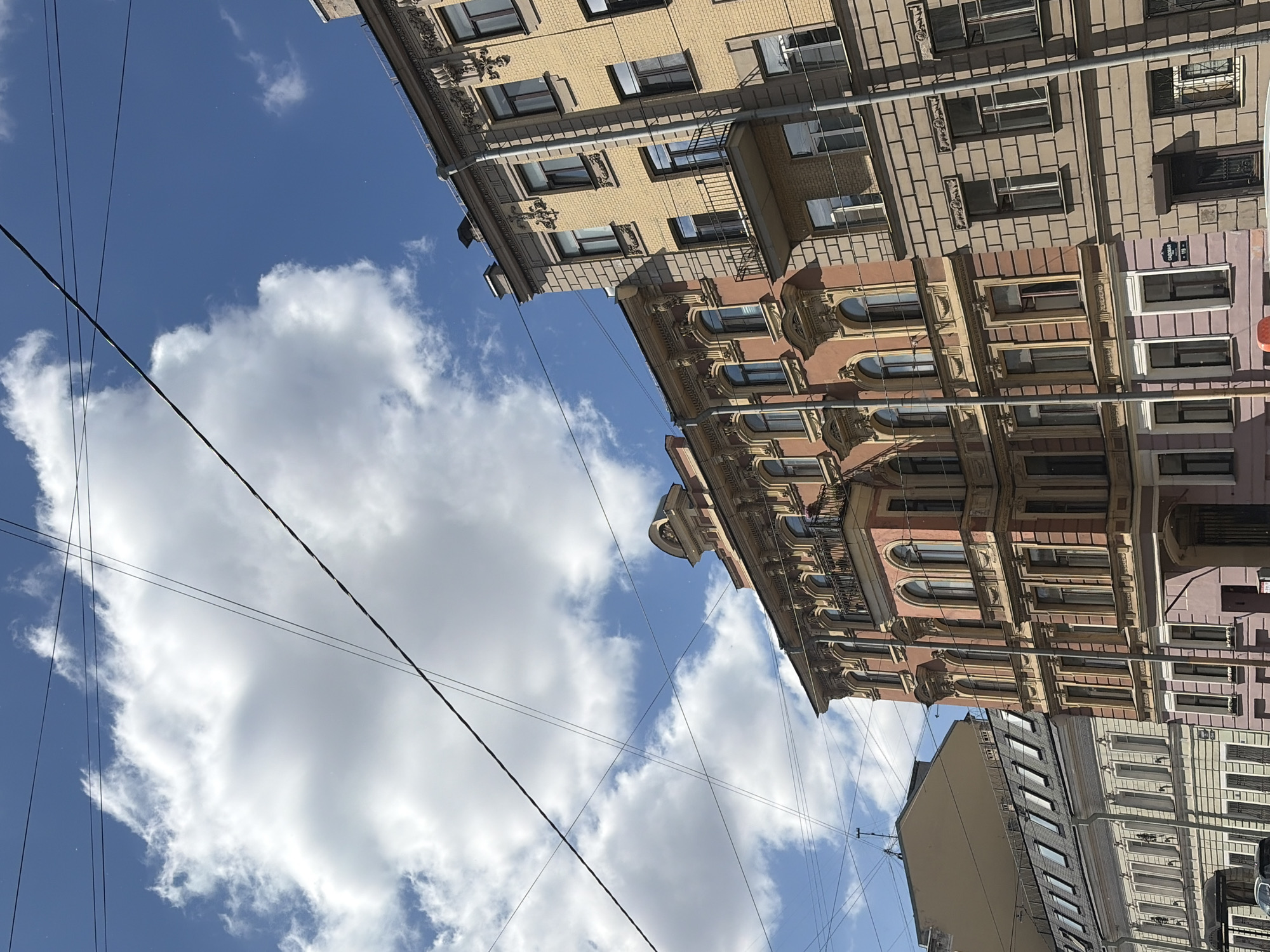 The image shows a view looking up at buildings in Saint Petersburg, Russia, under a partly cloudy blue sky. Multiple overhead wires crisscross the sky. The buildings appear to be old, with ornate architectural details. The building in the foreground on the right is yellow brick, with a decorated facade. To its left is a pinkish-red building with similar decorative elements and arched windows. Further to the left, another building with a mansard roof is visible, followed by a lighter-colored building with numerous windows. No people are clearly visible in the frame. The overall scene depicts an urban streetscape with classic European architecture under a bright, sunny sky.