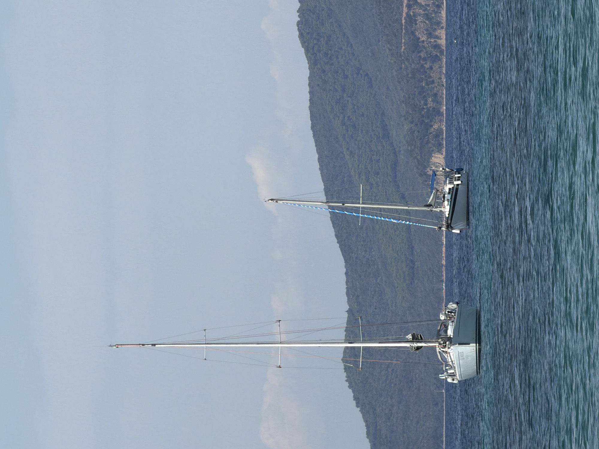 Two sailboats are anchored in the clear blue waters off the coast of Punta Ala, Italy, with a lush green, tree-covered mountain range forming the backdrop. The sailboat on the left is larger and closer to the viewer, its mast rising prominently against the pale blue sky. The second sailboat, smaller and further to the right, displays a blue and white striped sail or flag. The water is calm with gentle ripples, reflecting the bright sky. The scene appears to be a peaceful, sunny day.