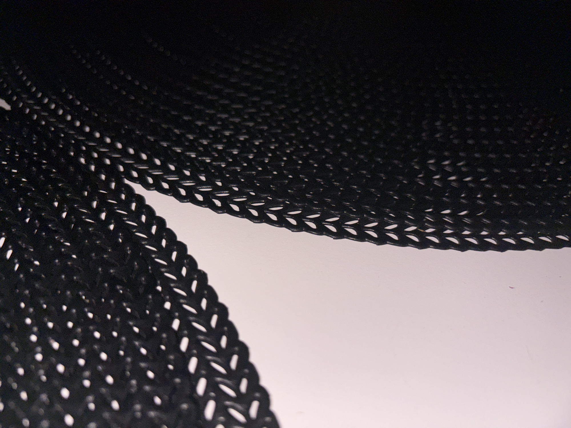 This is a close-up, abstract shot of a black, woven material that appears to be synthetic, possibly nylon or plastic. The material has a repetitive, chain-like pattern with small oval-shaped openings. It is illuminated from the side, creating a shadow effect and highlighting the texture and depth of the weave. The background is a soft, gradient of white to pale pink, suggesting a diffused light source or a colored surface. The image has a shallow depth of field, with the foreground material in sharp focus and the background softly blurred. There are no people or clear objects identifiable in the scene, making it difficult to place it in a specific location within Cupra Marittima, Italy. The context of the image is purely textural and abstract, focusing on the material's properties and the interplay of light and shadow.