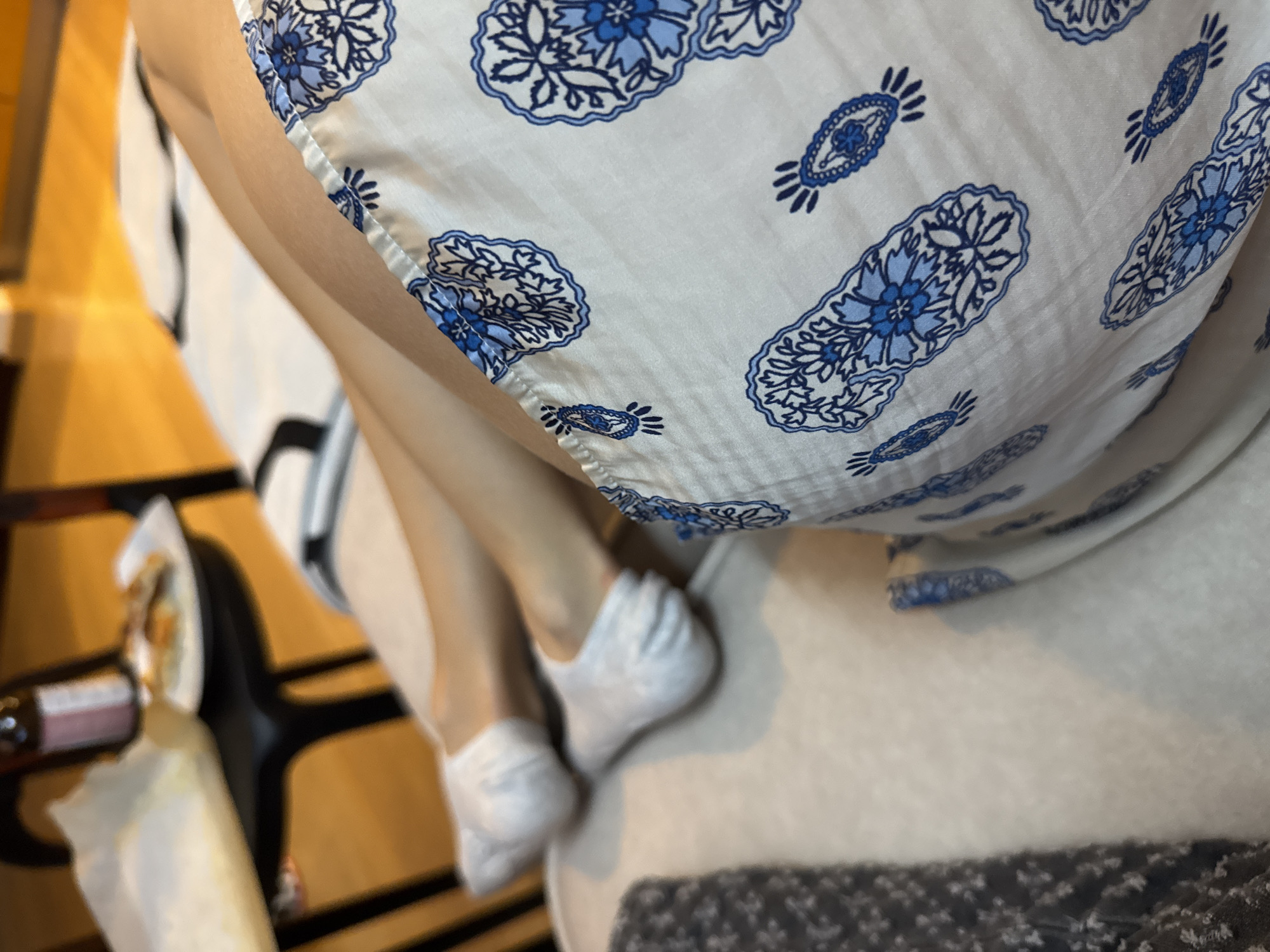 The image presents an overhead view of a person relaxing indoors, with the focus on their lower body. The person is wearing a white garment featuring a distinctive blue floral and paisley-like pattern. Their legs are bent, and their feet, covered in white, soft-looking socks or foot coverings, rest on a light-colored surface, possibly a bed or couch.
To the left, a dark, blurry surface, likely a table or tray, holds various items including a bottle (possibly containing a beverage), a plate with some food, and food wrapped in paper. This suggests a casual setting where the person might be enjoying a snack or meal. In the bottom right corner, a darker, patterned fabric, perhaps a blanket or rug, is partially visible. The lighting indicates an indoor environment, possibly illuminated by artificial light. The scene conveys a sense of comfort and leisure. No legible text is discernible on any of the objects.