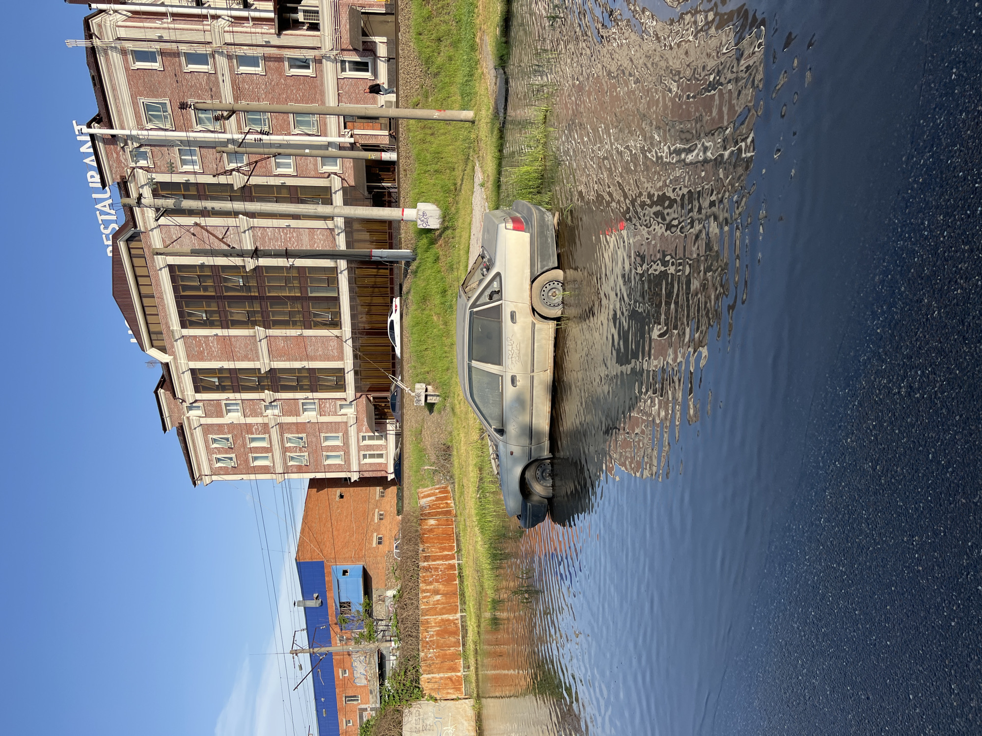 A dilapidated gray car sits partially submerged in a flooded area near a building in Krasnodar, Russia. The building has a restaurant. The water reflects the building and the sky. A section of pavement is visible.