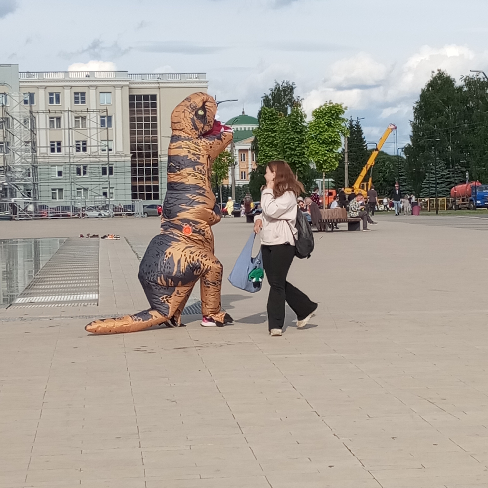 This image depicts a daytime scene in Central Square, Izhevsk, Russia. In the foreground, an individual in an inflatable T-Rex dinosaur costume, featuring a brown and black camouflage pattern and a visible fan unit on its left hip, is positioned in a kneeling or crouching posture, facing to the right. A woman, with reddish-brown hair, is walking from right to left past the dinosaur. She wears a light-colored hoodie, dark pants, and light-colored sneakers, carrying a dark backpack on her left shoulder and a light blue fabric bag containing a green object in her right hand. Her head is turned towards the dinosaur or slightly beyond it, with her right hand near her chin.
The ground is a light-colored paved surface with visible seams. To the left, a water feature or architectural element with parallel lines is visible. In the midground, benches with several individuals sitting are present.
The background features several buildings and trees under a partly cloudy sky. Prominently, the Izhevsk State Circus building with its distinctive green dome is visible behind the dinosaur. To the far left, a multi-story building is partially obscured by extensive scaffolding, suggesting construction or renovation. Further in the background, to the right, trees line the square, and various vehicles including a yellow crane, an orange truck, and a blue tanker truck are visible on a distant road. Additional people are walking or standing throughout the background.