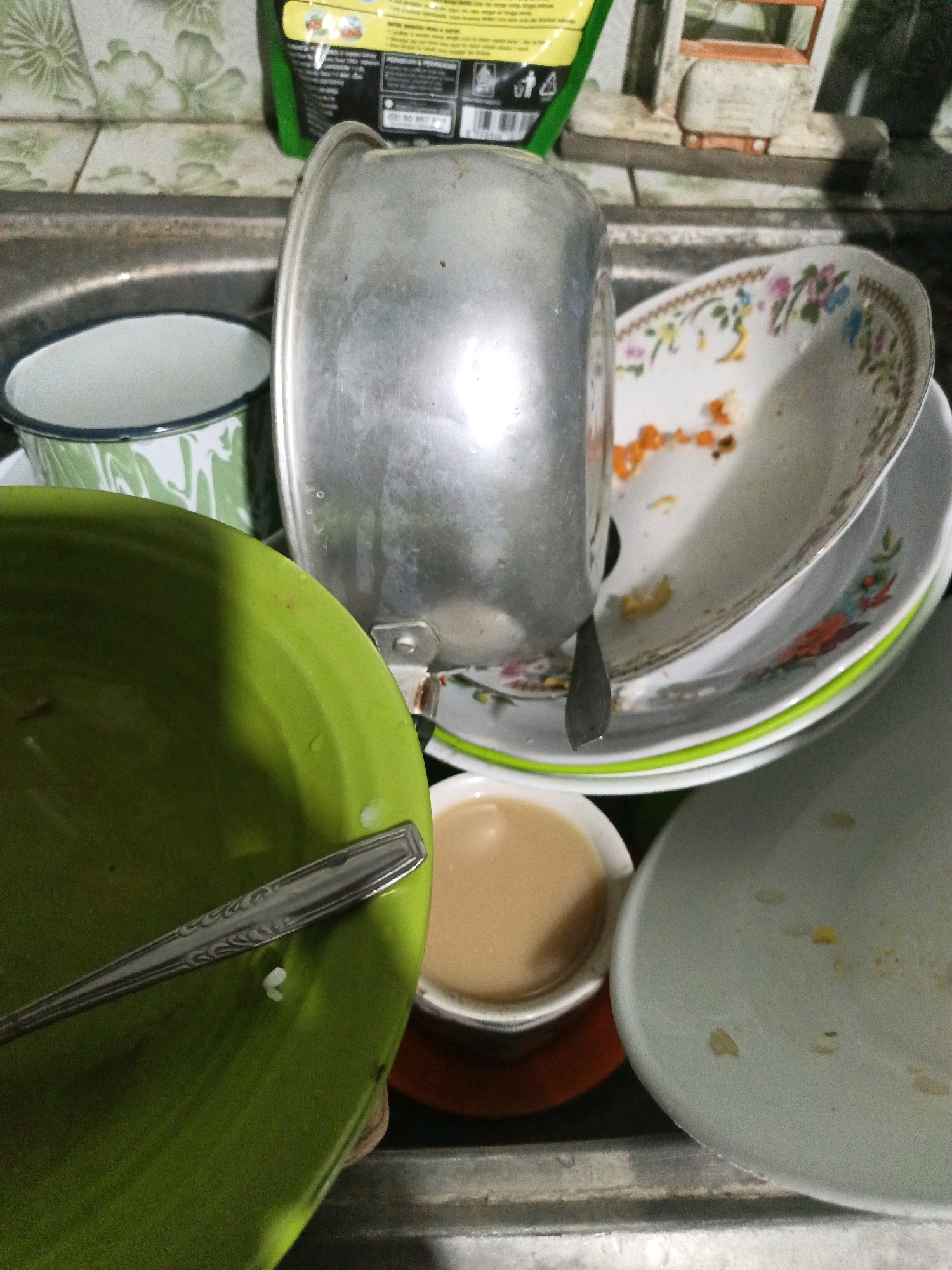 In Keleyan, Indonesia, a sink filled with dirty dishes presents a scene of domesticity. A large, dented aluminum pot dominates the center of the image, positioned precariously on top of a stack of plates. A green bowl, partially filled with water and a single spoon, sits to the left, its vibrant color contrasting with the muted tones of the surrounding clutter. To the right, a smaller, white bowl holds a light brown liquid, possibly tea or coffee, suggesting a recent meal or beverage. The sink itself, made of stainless steel, shows signs of wear and tear, with water stains and food residue visible. In the background, a green and white patterned mug peeks out from behind the aluminum pot. A partially visible bag of some sort, with yellow and green branding, hangs above the sink, adding to the overall impression of a lived-in, functional space. The scene is illuminated by natural light, casting shadows and highlighting the textures of the various objects.