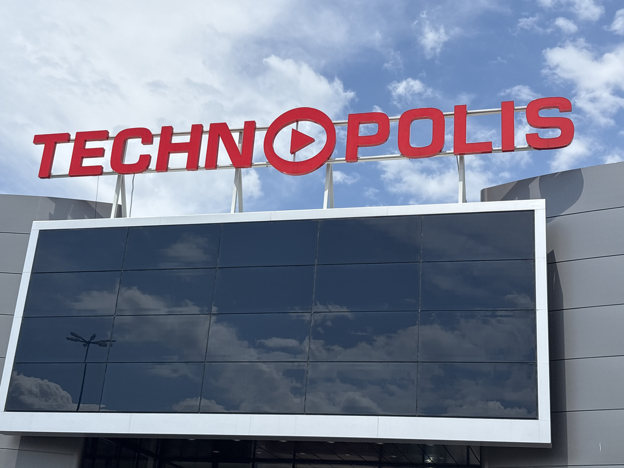 The image shows the Technopolis store in Plovdiv, Bulgaria. The red "TECHNOPOLIS" sign, with a play button symbol replacing the "O," is prominently displayed on top of the building. Below the sign is a rectangular structure with reflective windows that mirror the cloudy sky. A light pole is visible in the reflection. The building has gray siding. The sky is light blue with scattered white clouds.