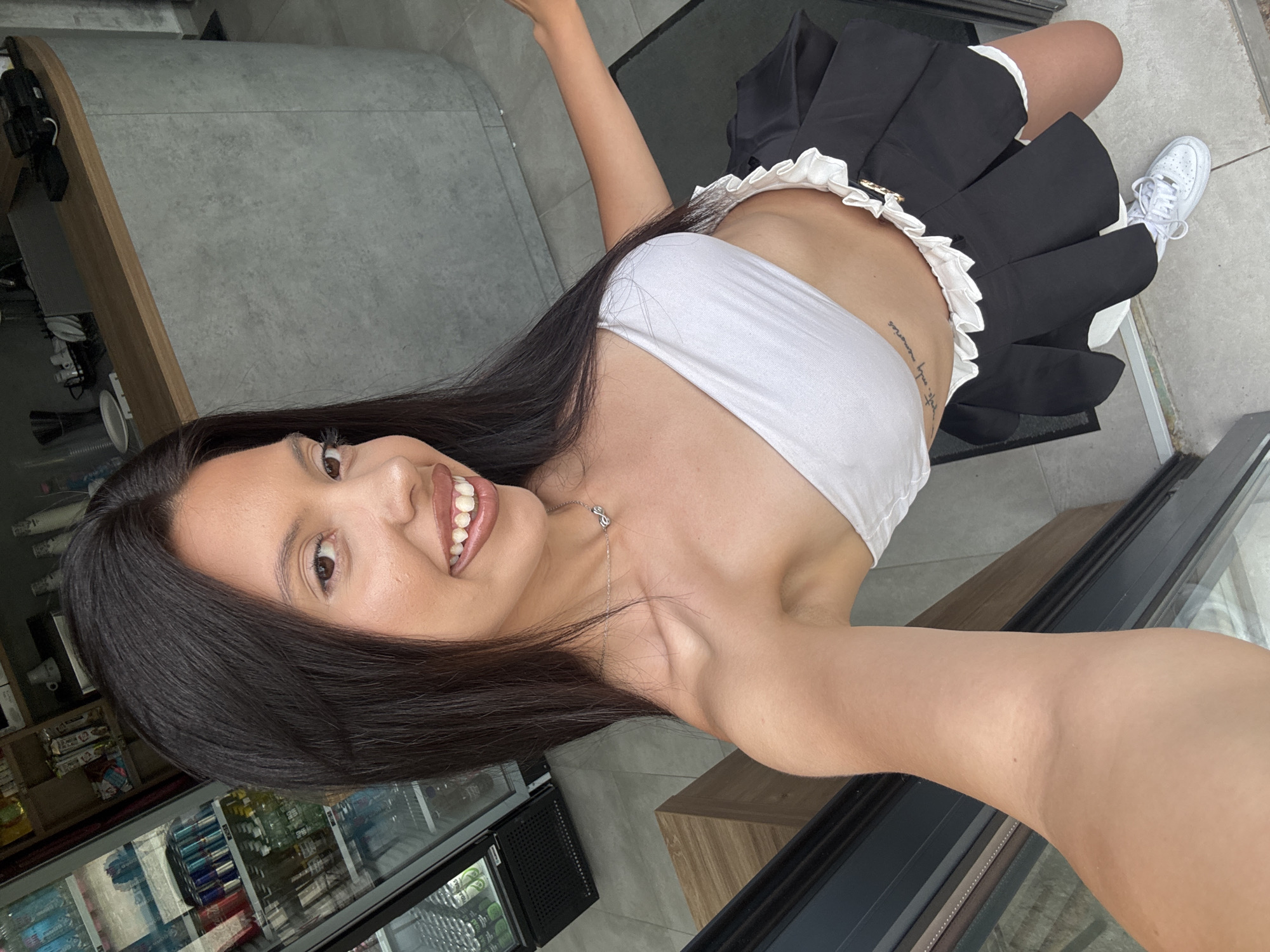 A young woman of apparent Mediterranean or Latin American descent, with long dark hair and brown eyes, smiles at the camera while taking a selfie. She is wearing a white tube top and a black and white pleated skirt, with white sneakers. She has a necklace with two intertwined loops. A tattoo is visible on her left side. The background appears to be an indoor-outdoor setting, possibly a cafe or shop, with a refrigerated display case filled with beverages on the left, and a counter with a gray curved surface on the right. The floor is tiled. The scene is lit with natural light. The location is the city of Sofia, Bulgaria.