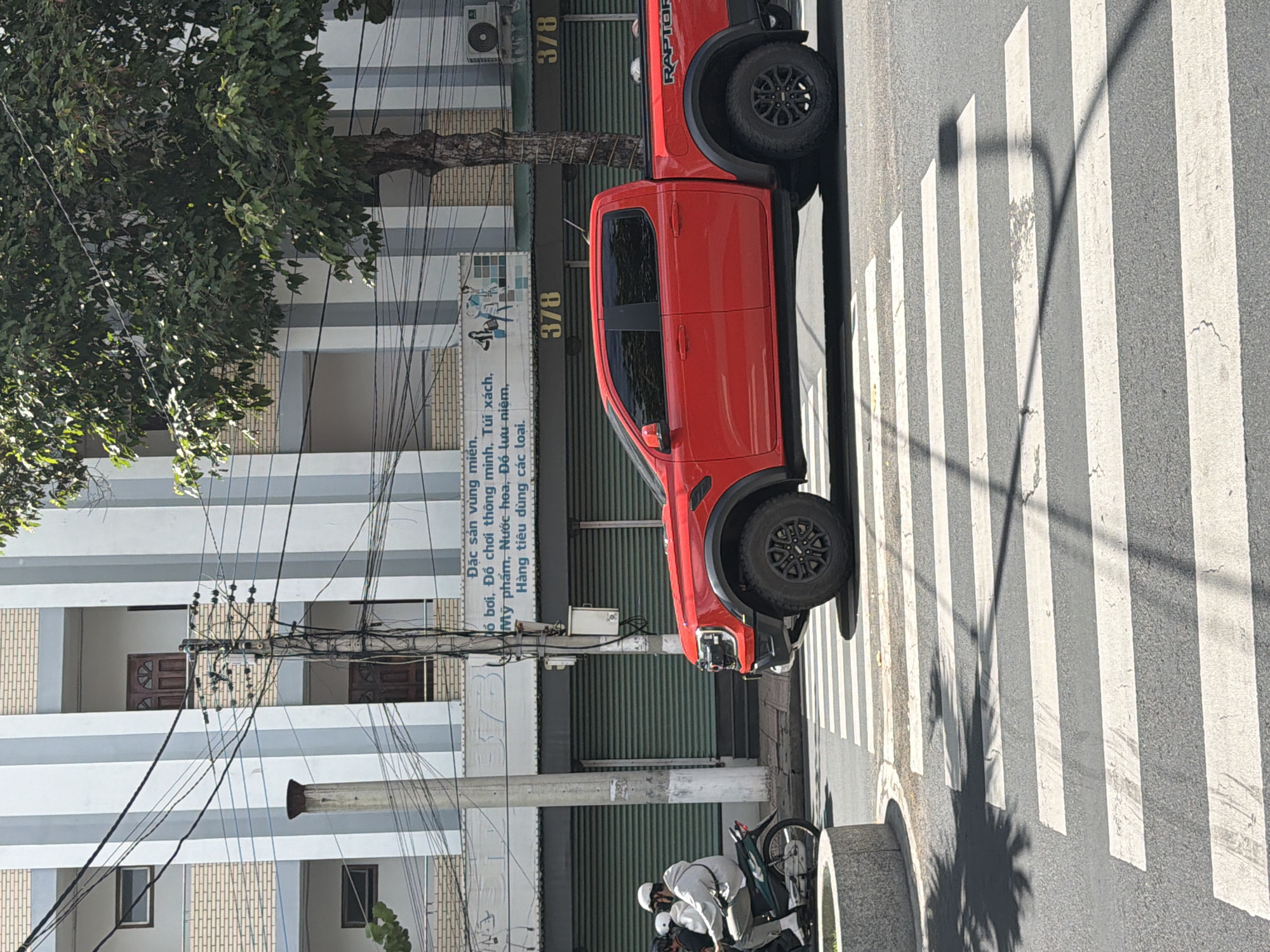 The image depicts a vibrant street scene in Nha Trang Ward, Vietnam, under bright, sunny conditions, indicated by strong shadows cast across the road. A striking red Ford Raptor pickup truck is a central subject, positioned on or very near a pedestrian crosswalk, facing right.
On the sidewalk to the left of the truck, two individuals in light-colored clothing and helmets are seen attending to a scooter. The background features multi-story buildings with balconies, windows, and closed roll-down shutters on their ground floors, implying commercial use. A large, green tree with a visible trunk partially obscures the upper left portion of the scene.
The urban infrastructure includes multiple utility poles densely covered with tangled wires, a street lamp, and concrete barriers or planters along the sidewalk. The road surface shows the distinct white stripes of a pedestrian crossing.
Visible text on a storefront includes Vietnamese phrases: "Đặc sản vùng miền, đồ bơi, Đồ chơi thông minh, Túi xách, Mỹ phẩm, Nước hoa, Đồ lưu niệm, Hàng tiêu dùng các loại." This translates to "Regional specialties, swimwear, smart toys, handbags, cosmetics, perfumes, souvenirs, all kinds of consumer goods." The number "378" appears twice on the building. The word "RAPTOR" is distinctly visible on the side of the red pickup truck's bed, confirming its model.