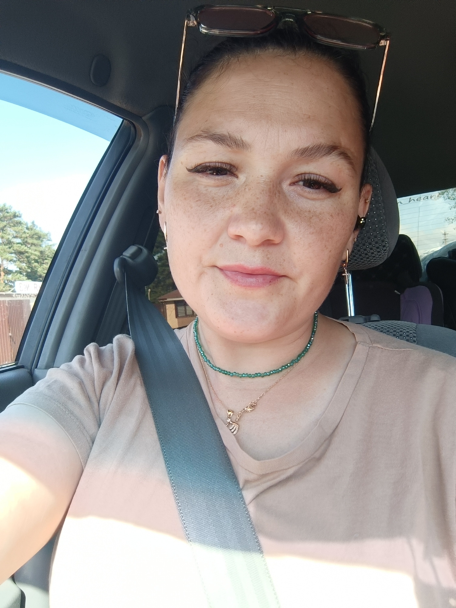 A medium shot selfie shows a woman with light skin and freckles sitting in the driver's seat of a car. She has dark hair pulled back and is wearing a light-colored t-shirt, a beaded green necklace, and a delicate silver necklace with a small pendant. She is also wearing a seatbelt. The background includes a car interior, a car window revealing trees and a building, and a headrest of a passenger seat. The image appears to have been taken on a sunny day. The photo was taken in Chaikovsky, Russia.