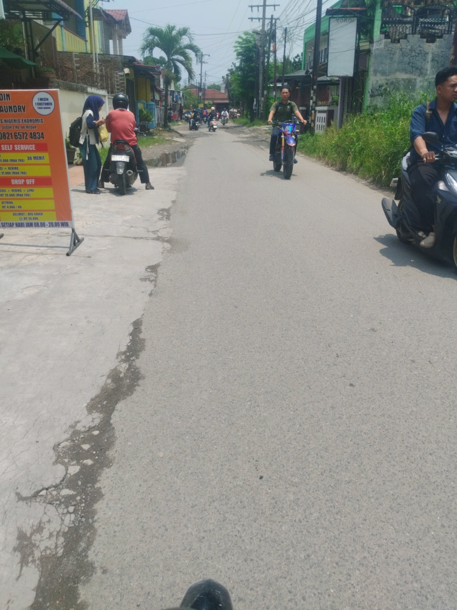 The image depicts a street scene in the city of Medan, Indonesia. On the left side of the image, a laundry service sign is visible, listing prices for various services. Next to the sign, a woman wearing a blue hijab and a white shirt stands by a motorcycle. A man in a red shirt is seated on the motorcycle. Further down the street, several other motorcycles are seen, with people riding them. The street is paved and appears to be a typical urban road. Buildings and vegetation line the street, indicating a residential or commercial area. The sky is clear, suggesting a sunny day. The overall scene captures a moment of daily life on a street in Medan.