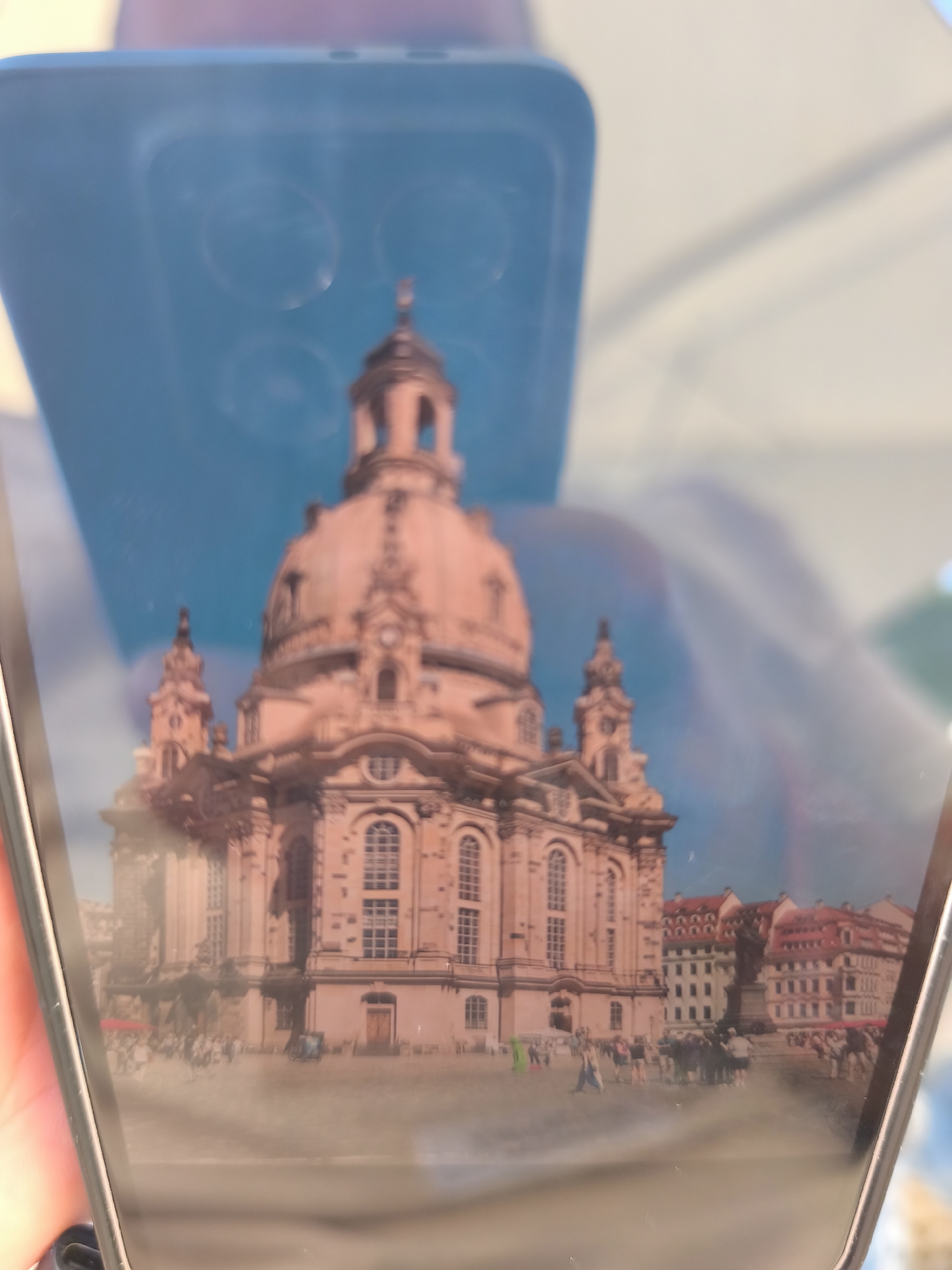 The image shows a reflection of the Frauenkirche Dresden in a phone screen. The church, with its distinctive dome, dominates the scene. A crowd of people is visible in the foreground, appearing to be in the square in front of the church. Other buildings can be seen behind the church. The phone itself is visible around the edges of the image. The location is likely in Dresden, Germany.