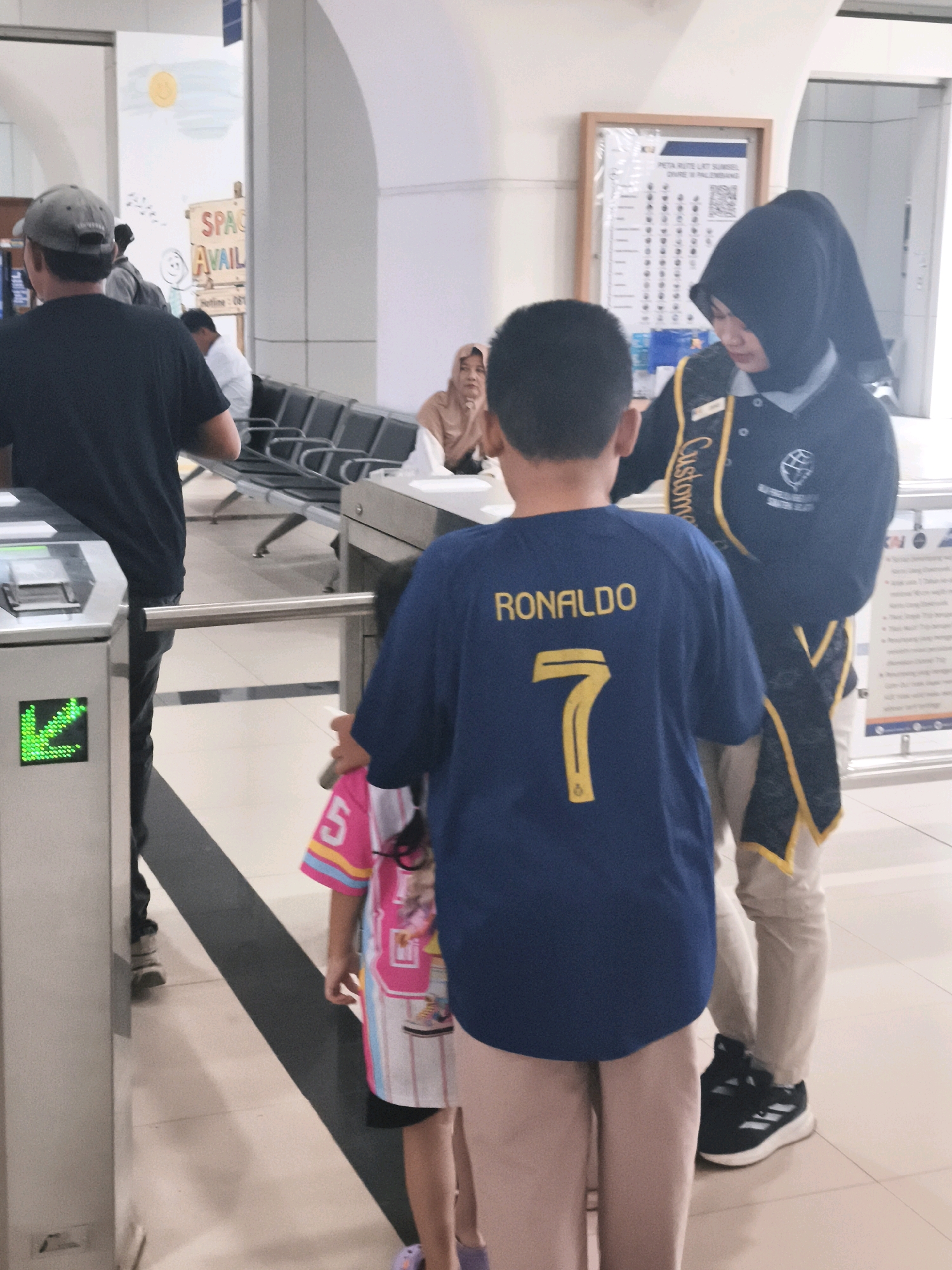 The image appears to be taken inside a modern building, possibly an airport or train station, in Kelurahan 16 Ilir, Kec. Ilir Timur I, Indonesia. In the foreground, a young boy wearing a blue t-shirt with "RONALDO" and the number "7" in yellow is visible from behind, facing away from the camera. To his right, a woman wearing a black hijab and a dark uniform with yellow trim, possibly a security or customer service employee, stands looking down. Next to the boy, a young girl in a pink and white patterned t-shirt with the number "5" is partially visible. They are standing near a metal turnstile with a green directional arrow illuminated on a digital display. In the mid-ground, a man wearing a black t-shirt and a grey baseball cap is seen from behind, walking away from the camera. Behind him, rows of empty seats are arranged, and a sign with "SPACE AVAIL" in colorful letters is visible on the wall. In the background, another person wearing a light brown hijab is seated. The environment is clean and well-lit, with tiled flooring and neutral-colored walls.