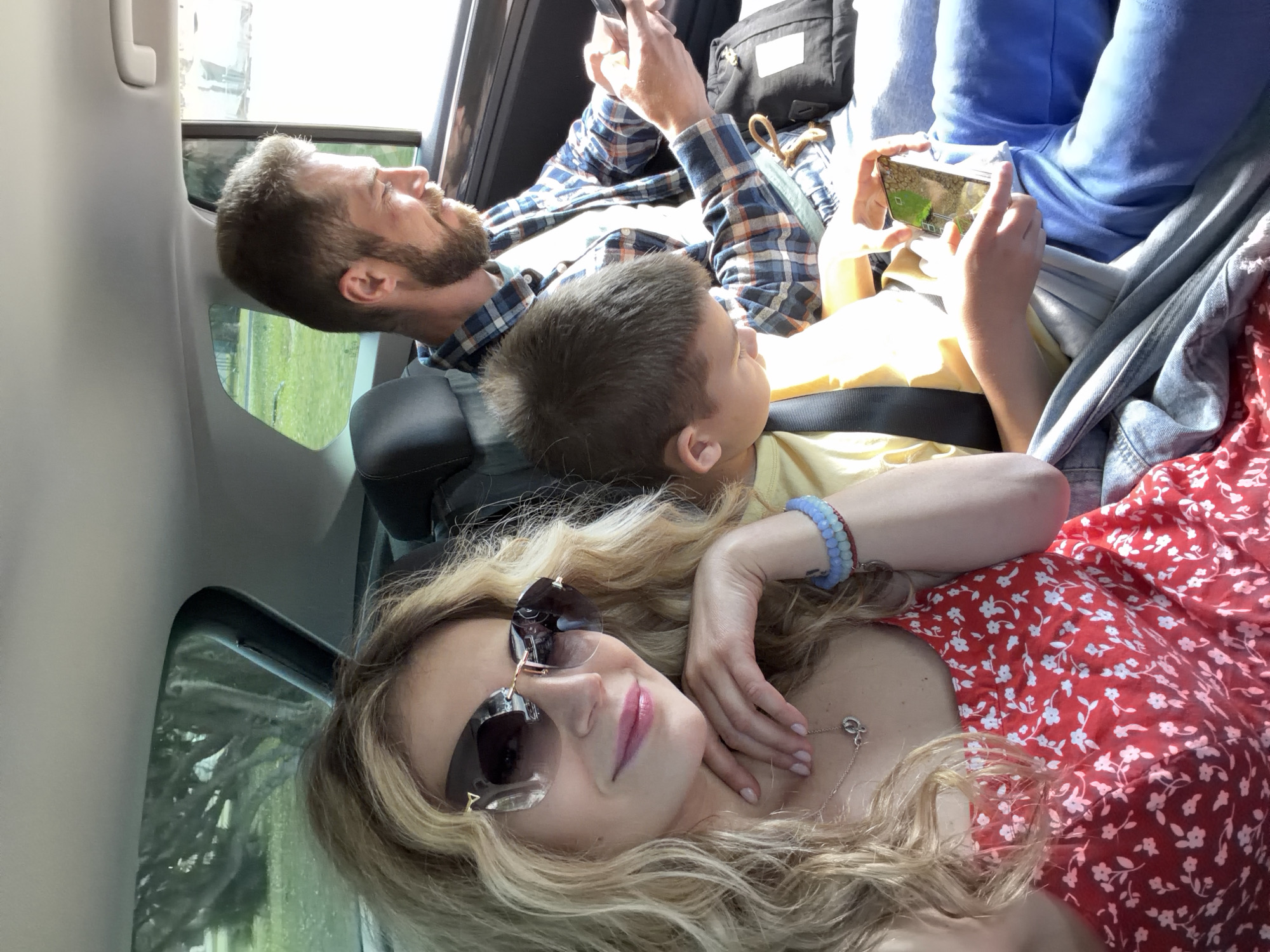 A family of three is in a car, lying in the back. The woman is in the lower left of the frame wearing sunglasses, a red floral dress, and a silver necklace. Her blonde hair is styled in loose waves. The boy is in the middle of the frame wearing a yellow shirt, looking up at a device being held by the father. The father, to the left of the boy, wears a blue plaid shirt and is also looking at a device. The background shows a glimpse of greenery and the car's interior. The location cannot be determined.
