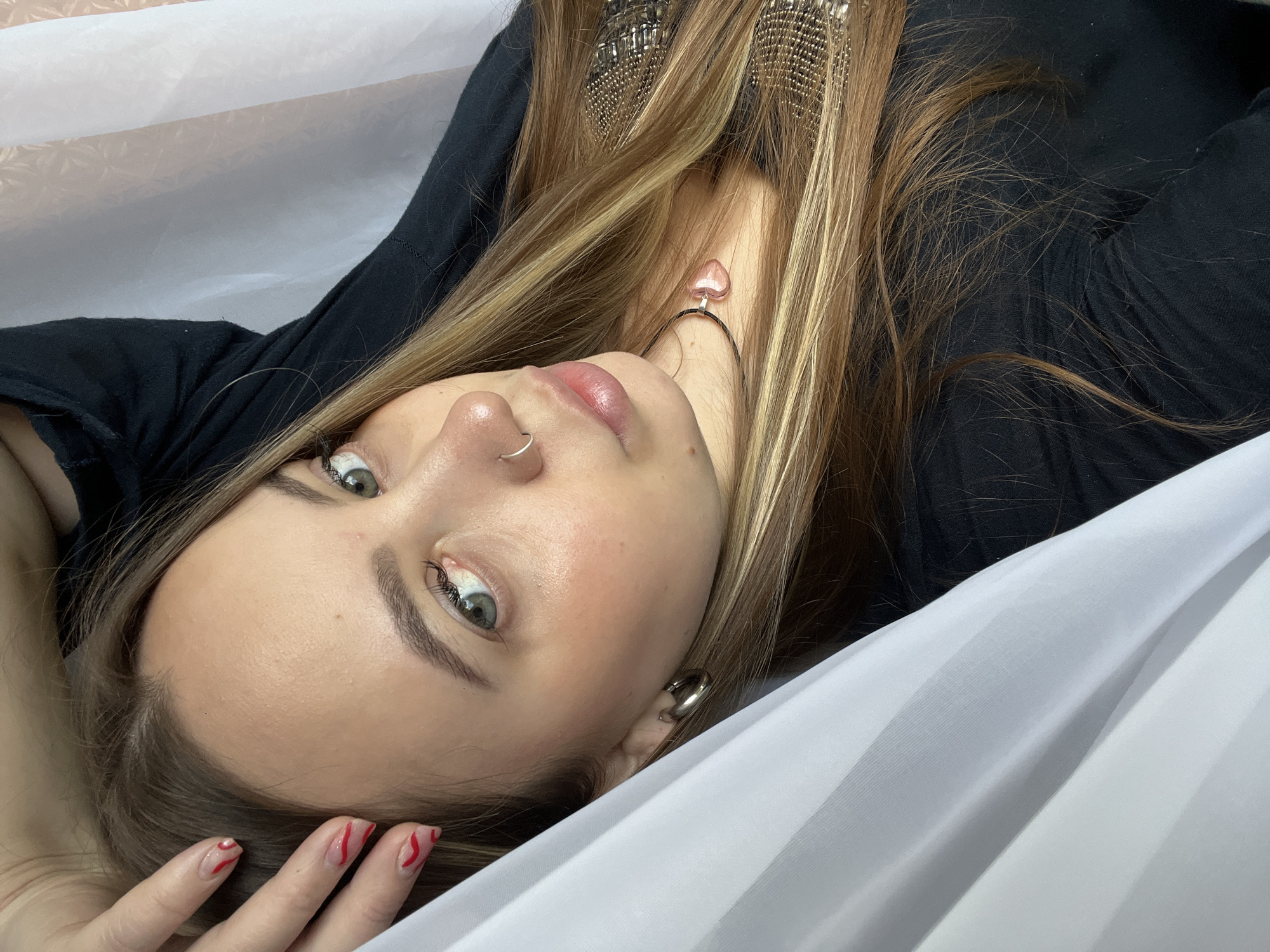 This is a close-up portrait of a young woman lying down, looking directly up at the camera. She has long, flowing light brown and blonde hair with prominent highlights. Her eyes are a light green or hazel, and she has mascara on her eyelashes with well-groomed eyebrows. A small silver hoop nose ring is visible on her left nostril. Her lips appear full and naturally pink, possibly with a subtle gloss.
She is wearing a black top or shirt. Several pieces of jewelry are visible: large, intricate metallic earrings or a headpiece on her right side, a necklace with a distinctive pink, heart-shaped pendant, and a silver hoop earring on her right earlobe. Her fingernails feature a clear base with stylized red wavy or zig-zag lines.
The woman is positioned against a background of draped white fabric, which creates a soft, luminous setting. She appears to be posing for the photograph. The image provides no specific indicators for the time of day or weather conditions. Based on the provided context, the location is Novopokrovskaya, Russia. There is no visible text in the image.