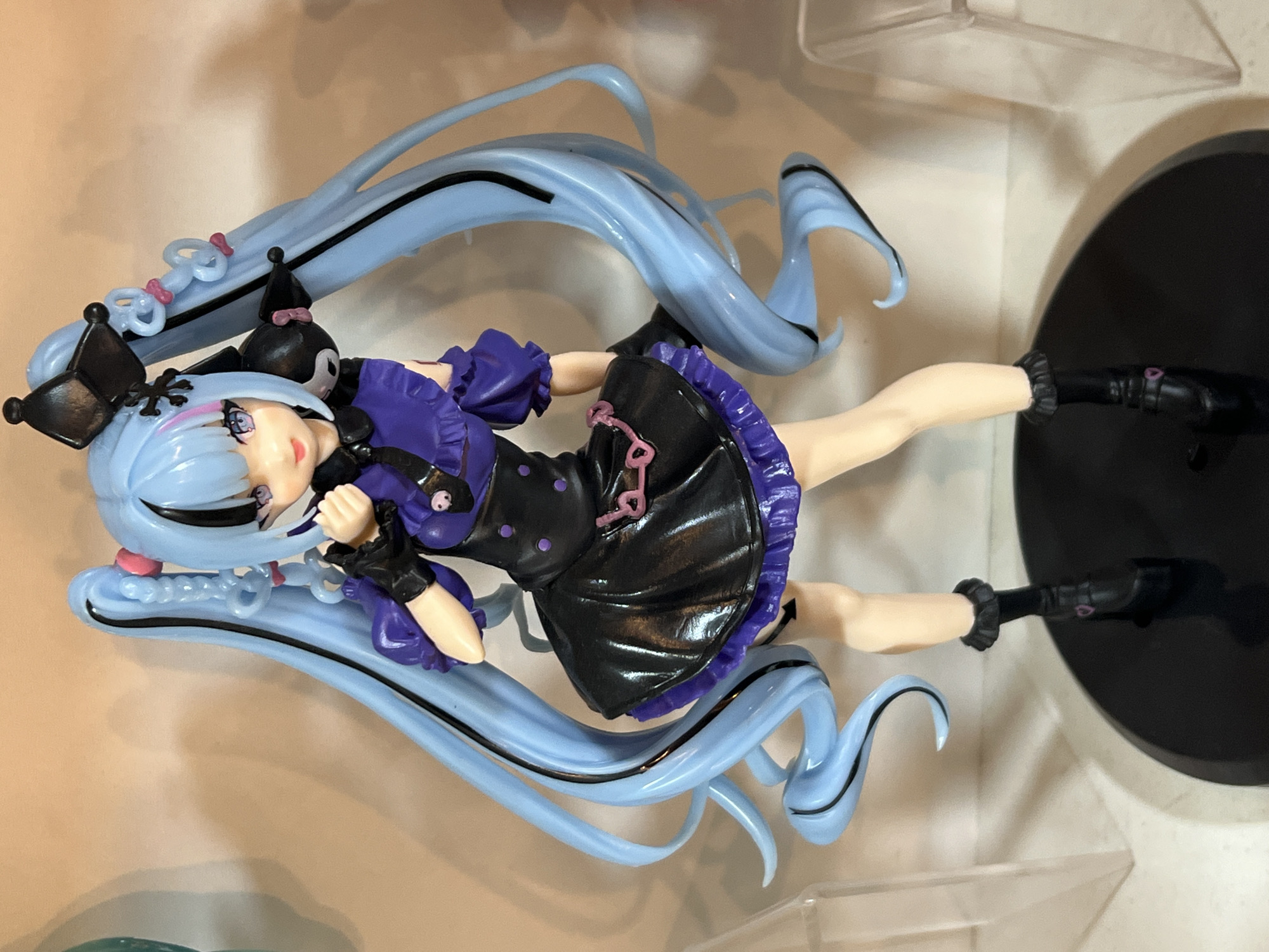 A full shot of an anime figurine shows a female character with long, light blue hair, styled in pigtails and flowing down her back. She has purple eye shadow and dark lipstick. She is wearing a black dress with a purple ruffled trim and a matching purple top with ruffled sleeves. The figurine is standing on a black circular base. The background is a plain, light-colored wall. The scene does not depict any identifiable urban or natural environment, nor does it provide any clues to a specific geographical location such as Arica, Chile.