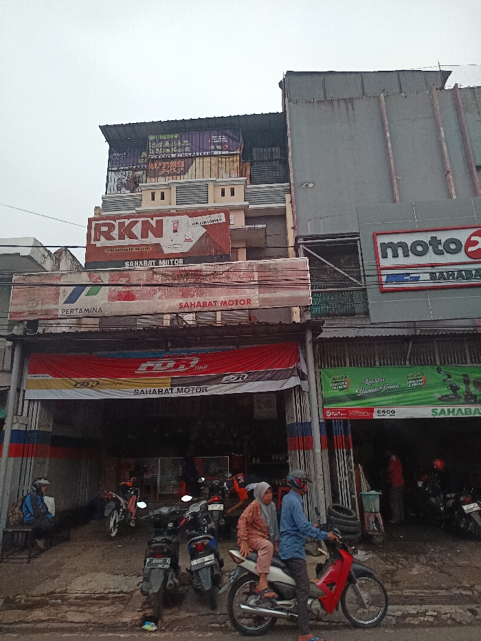 This image presents a street-level view of a commercial district in Ciranjang, Indonesia, under an overcast sky. The scene is dominated by two multi-story buildings housing motorcycle-related businesses.
The building on the left features a ground-floor entrance covered by a red, yellow, and white banner displaying "FDR TIRE" and "SAHABAT MOTOR." Above this, a weathered horizontal sign reads "PERTAMINA SAHABAT MOTOR," beneath a larger "RKN SAHABAT MOTOR" sign with decorative architectural elements. The upper floors are clad with various large, faded banners and corrugated metal sheeting.
To its right, an adjacent multi-story building displays a grey, vertically ribbed facade. Its ground floor hosts another business, indicated by a prominent green banner with white text, partially reading "JUARA" and "SAHABAT MOTOR." Higher on this building, a "motoX" sign is visible.
In the immediate foreground, on the uneven and damp dirt ground, a red and black motorcycle is occupied by two individuals. The driver, a male wearing a blue shirt and a grey helmet, sits astride the motorcycle. A female passenger, wearing a light-colored headscarf and a patterned brown top, sits side-saddle behind him. Multiple other motorcycles are parked or positioned within the open entrances of both shops. To the far left, a person wearing a blue jacket and a helmet is seated on a low wooden bench. Stacked motorcycle tires are visible near the entrance of the right-hand shop. Overhead, a dense network of electrical wires spans the street.