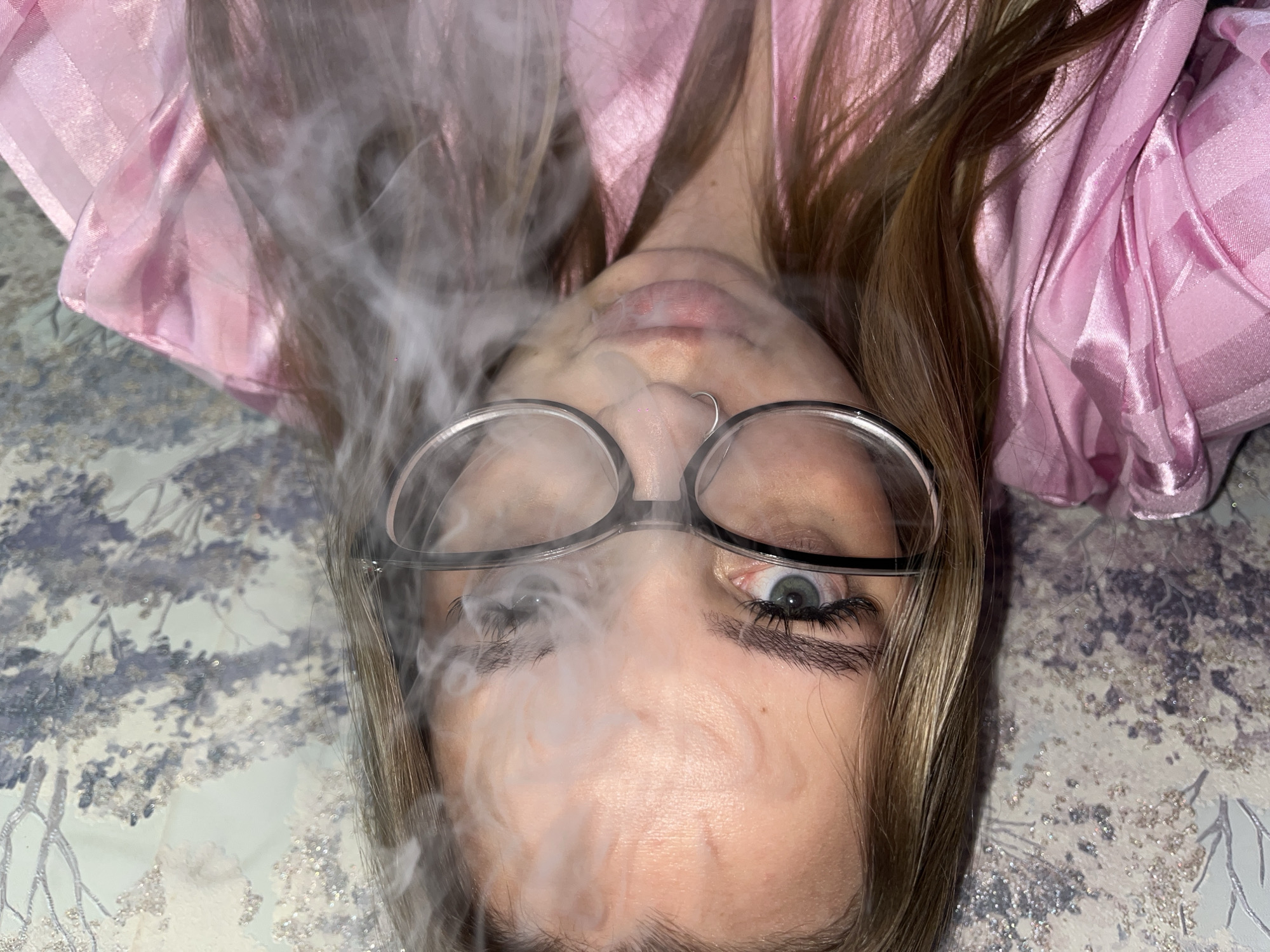 A woman with blonde hair and wearing glasses lies on her back, exhaling smoke. She is wearing a pink top. The background is a patterned surface. The image was taken in Novopokrovskaya, Russia.