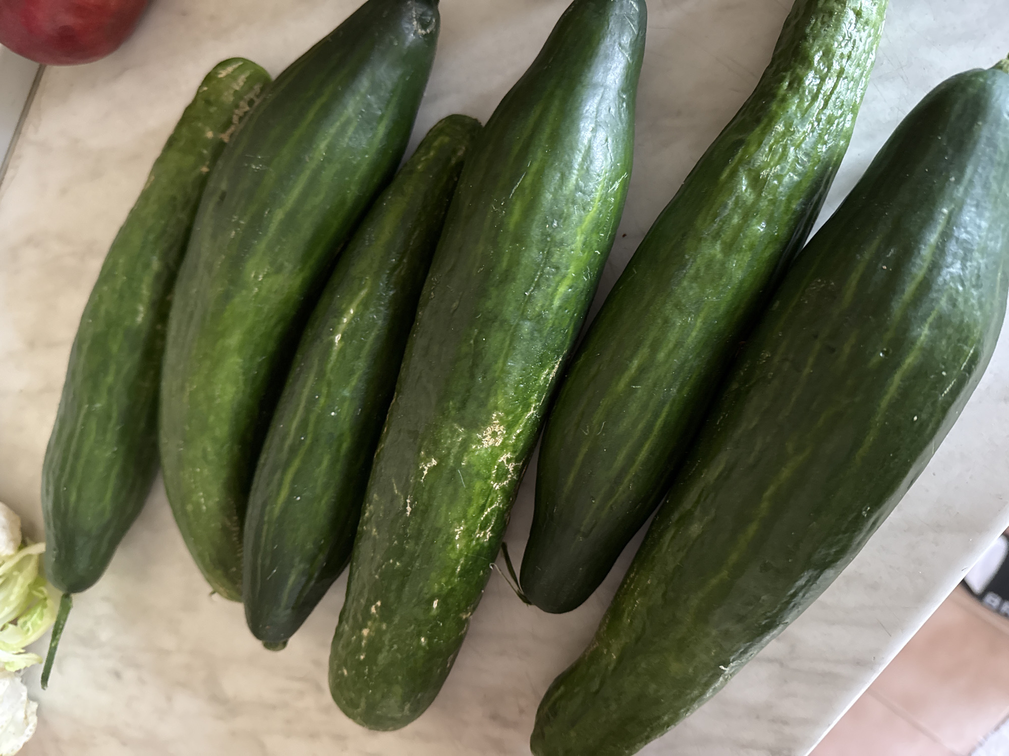 The image shows a close-up view of six green cucumbers lying on a white marble countertop. The cucumbers are of varying sizes and have dark green skin. A red fruit, possibly an apple, is visible in the upper left corner. A few leaves of a head of lettuce are in the lower left corner. The photo appears to be taken indoors.