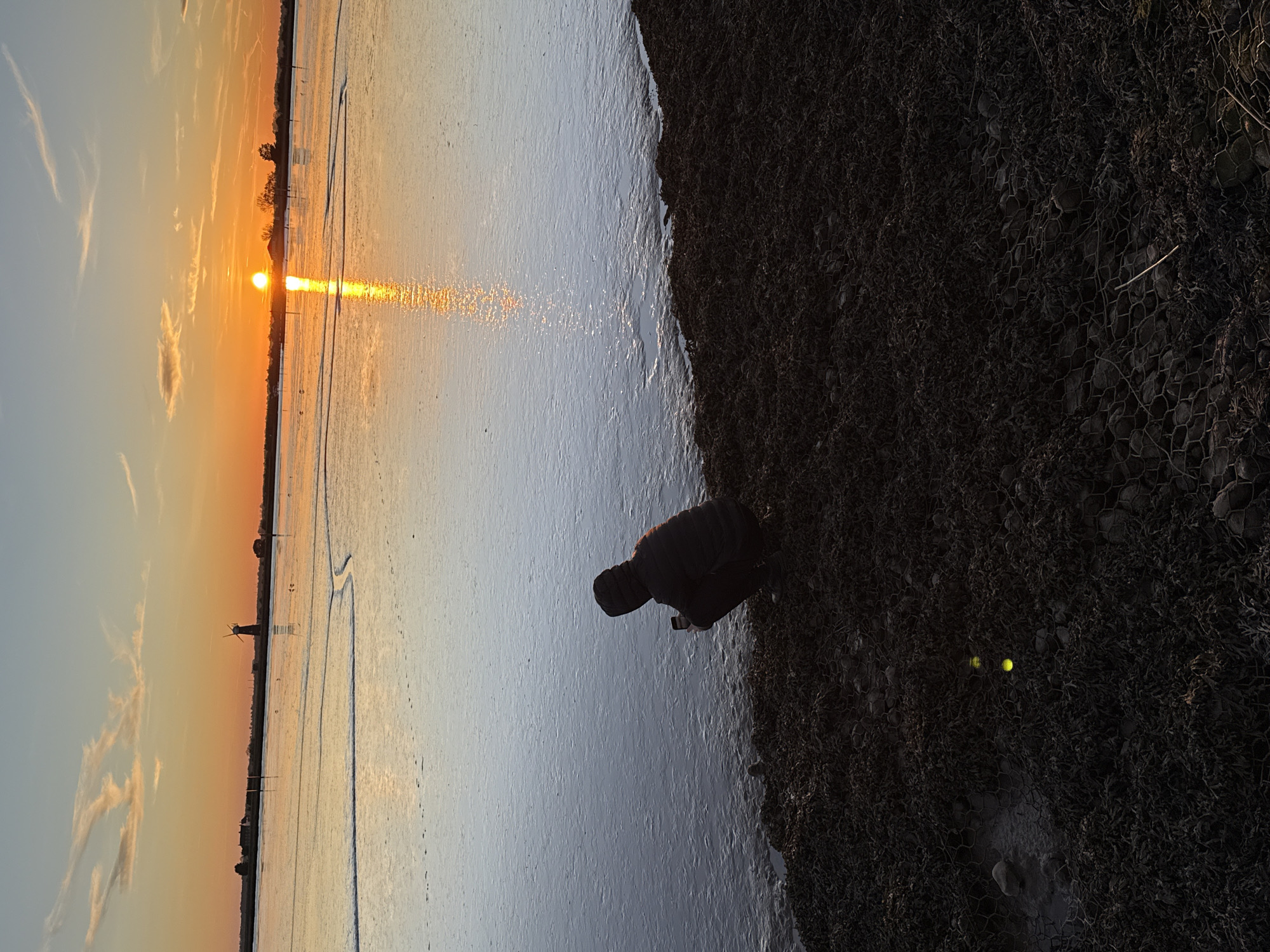 The image shows a coastal landscape at sunset in Great Yarmouth, United Kingdom. The setting sun casts an orange glow across the sky and reflects off the water. A person stands on a dark, textured ground near the water's edge. A silhouette of a windmill is visible on the horizon.