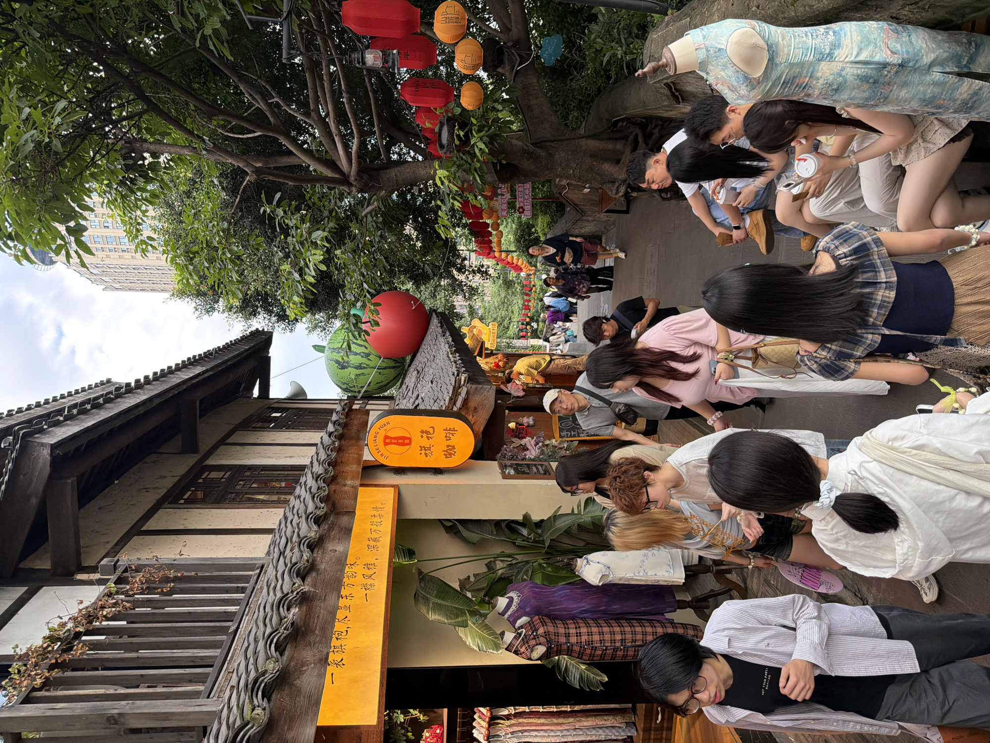 The image depicts a bustling pedestrian street in Yuzhong District, China, likely a tourist area given the traditional architecture and decorative elements. A narrow, paved pathway winds uphill, flanked by buildings with dark tiled roofs and white walls, suggestive of historical Chinese architecture. The buildings feature wooden balconies and window frames, with some displaying clothing on mannequins and hanging racks. Several shops are visible, one with an orange sign bearing Chinese characters.
The street is populated by a diverse group of people. In the foreground, several individuals are walking and observing the shops. A person in a white hooded jacket with dark hair pulled into a ponytail is facing away from the camera, holding a phone. To their left, a person with glasses and a striped shirt walks by. Further up the street, a group of people are seated on steps, some holding drinks. In the background, more pedestrians are seen walking, with a tall, modern building visible in the distance, contrasting with the older architecture.
Decorative elements are abundant. Red and orange lanterns are strung from trees and light fixtures, adding a festive atmosphere. A large, stylized watermelon and a red apple are suspended as oversized decorations above a shop. Lush green trees and foliage are interspersed throughout the scene, providing shade and natural beauty. The overall impression is a vibrant and lively streetscape, blending traditional aesthetics with contemporary life.