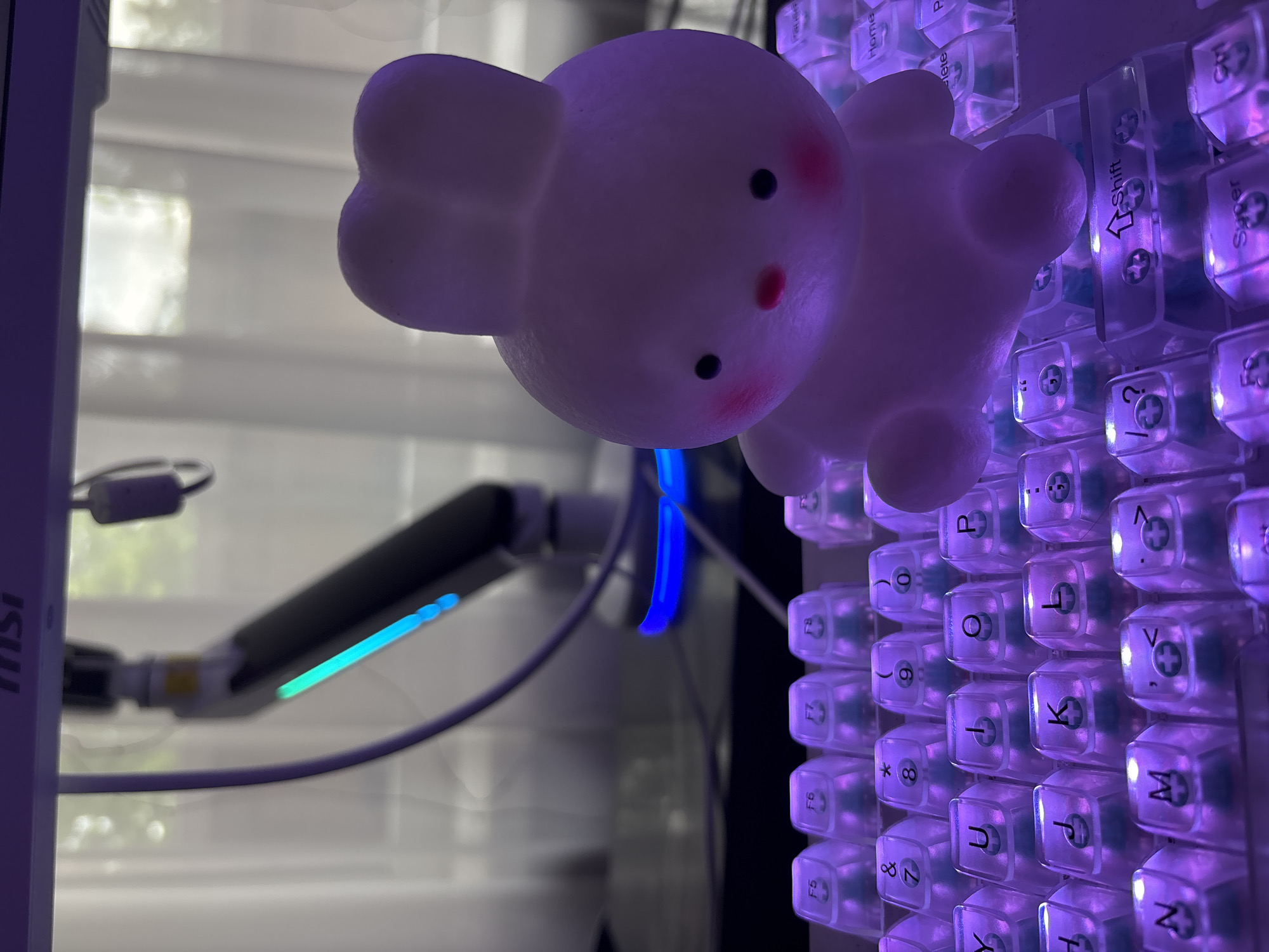 An illuminated bunny figurine sits on a purple-lit keyboard. The bunny, made of translucent material and emitting a soft glow, has small black eyes and rosy cheeks. It is positioned on the right side of the keyboard, facing slightly to the left. The keyboard features clear keycaps with purple backlighting, with various keys such as "F5," "F6," "F7," "F8," "Y," "U," "I," "O," "P," "H," "J," "K," "L," "N," "M," and "Shift" visible. In the background, a monitor arm with blue and green LED lights is visible, as well as sheer curtains and a blurred window. The lighting suggests an indoor setting, possibly a home office or gaming setup.