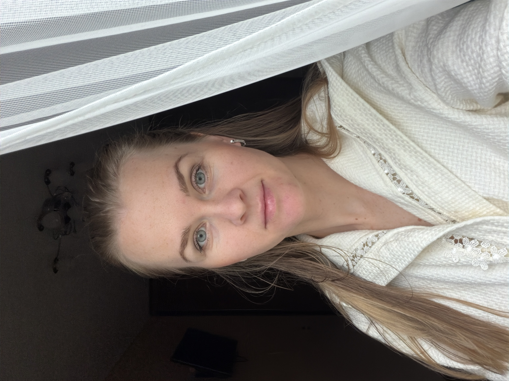 A young woman with blonde hair and blue eyes is pictured in a close-up selfie. She is wearing a white terrycloth robe with lace trim around the collar and pockets. Her hair is wet and down, falling over her shoulders. She is looking directly at the camera with a neutral expression. The background is dark and out of focus, with a hint of a curtain on the right side of the image. The lighting suggests an indoor setting, possibly a bathroom or bedroom. There is no discernible connection to the city of Khabarovsk, Russia.