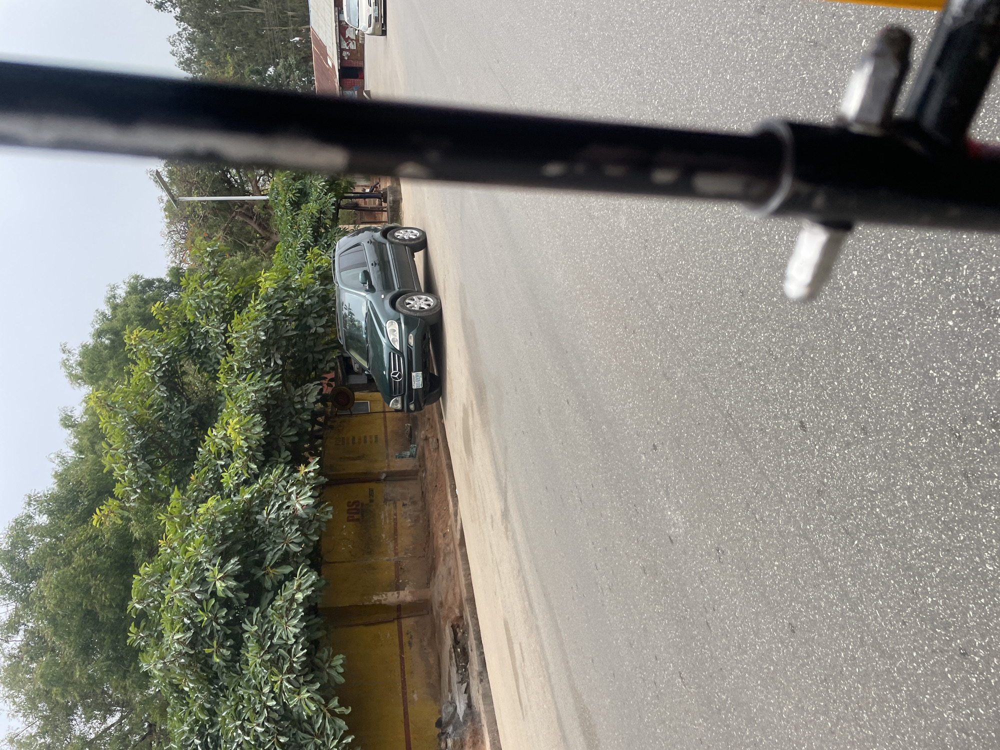 The image displays an outdoor street scene on a bright, clear day in what appears to be Jos, Nigeria. The photograph is taken from inside a vehicle, with a prominent black bar, likely part of the vehicle's frame, extending horizontally across the top portion of the image.
The primary subject is a dark-colored SUV, identified as a Mercedes-Benz M-Class (W163 generation), parked on the side of a paved road. Its license plate is partially visible, showing "LAGOS STATE" and the numbers "8833". The road surface is gray asphalt, appearing well-maintained.
Adjacent to the road and the parked SUV is a light brown, unpaved shoulder or sidewalk, which borders a low, ochre-yellow wall or building. This wall is largely covered by an abundance of lush, dense green bushes and trees, suggesting a verdant environment. Faint text and markings are visible on the yellow wall, including the clear letters "POS". Beyond these bushes, more trees with lighter green foliage rise against a bright, hazy sky.
No individuals or specific activities are clearly discernible in the frame, implying a quiet street. The scene is illuminated by daylight, with soft shadows, indicating it is likely midday or early afternoon.