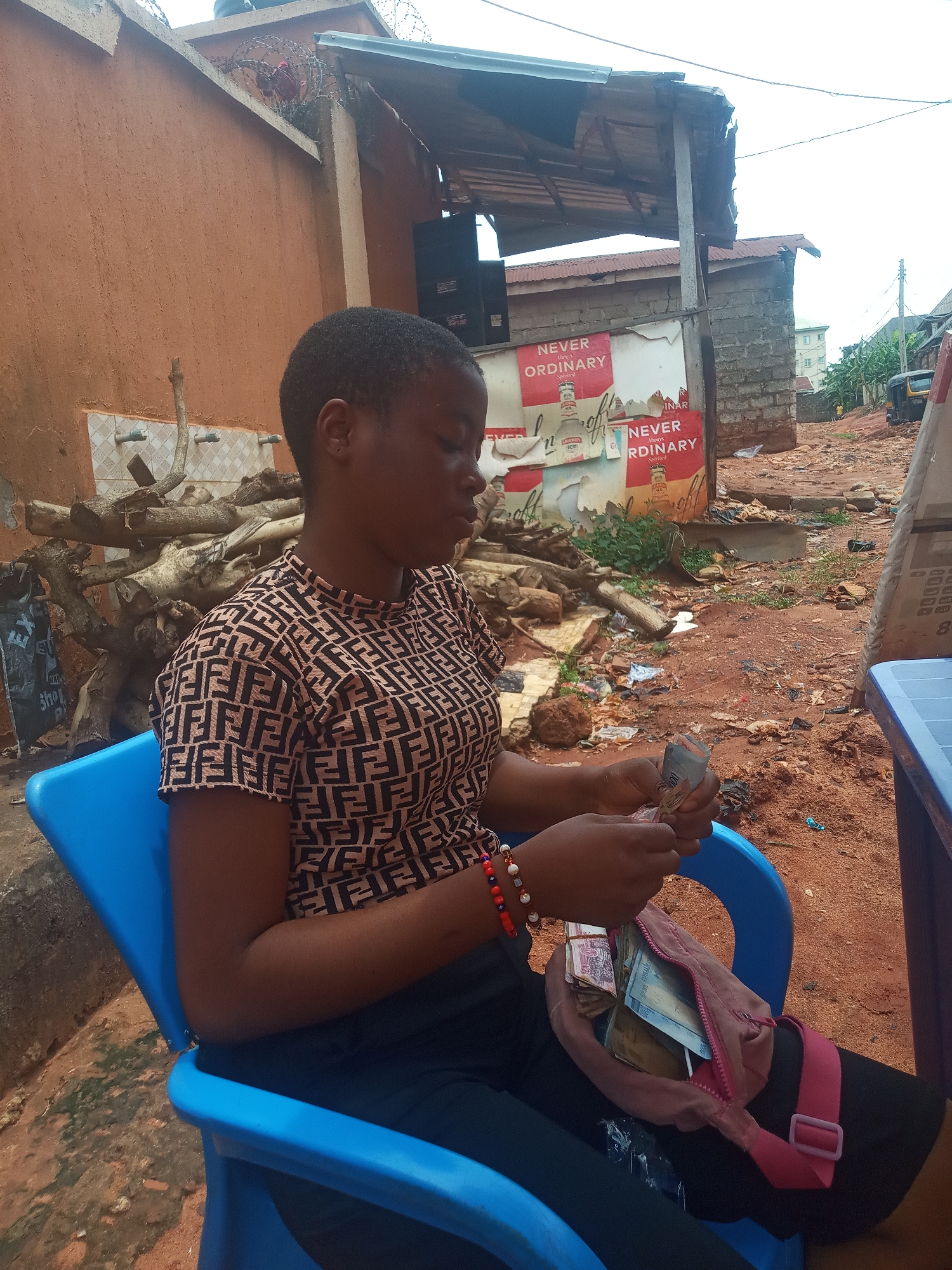 A young woman is seated on a blue plastic chair outdoors, likely in Ukwu-Abwa, Nigeria. She is counting a stack of Nigerian Naira bills. The currency is partially held in a pink fanny pack resting on her lap. She wears a patterned brown and black shirt, dark pants, and a colorful beaded bracelet. Background elements include a pile of logs, a partially visible advertising sign with the words "NEVER ORDINARY", and a rough dirt road. Nearby are buildings constructed of cinder block and corrugated metal.