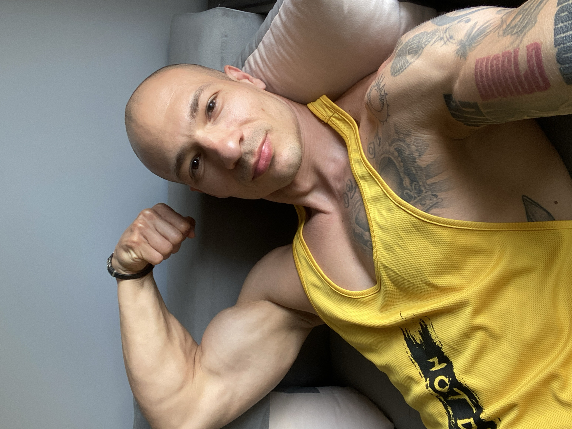 A bald, light-skinned man is reclining on a gray upholstered surface with a white pillow behind his head. He is wearing a yellow tank top with black writing partially visible on the front. He is flexing his left bicep, which is visible in the foreground. Tattoos are visible on his upper left arm and chest. The background is a plain gray wall. There is no evidence to indicate the city of Pleven, Bulgaria.