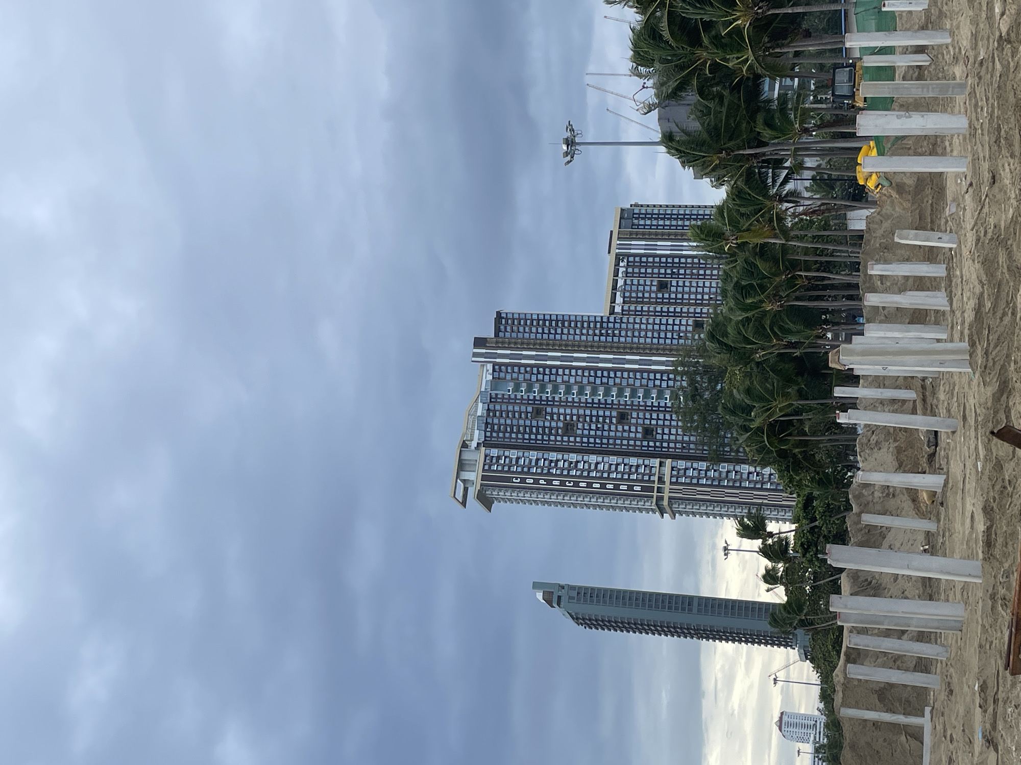 The image depicts a beach construction site in Pattaya City, Thailand, under a cloudy sky. In the foreground, numerous concrete pillars are erected on the sandy ground, indicating ongoing foundation work for a new structure. Behind the construction site, a row of palm trees sways in the wind, separating the beach from a backdrop of modern, high-rise buildings. Two prominent skyscrapers, one taller and sleeker, the other wider and more complex, dominate the middle ground. Further in the distance, other buildings and construction cranes are visible. A street lamp with multiple lights stands tall to the right of the skyscrapers. The overall scene suggests a developing urban coastal area.