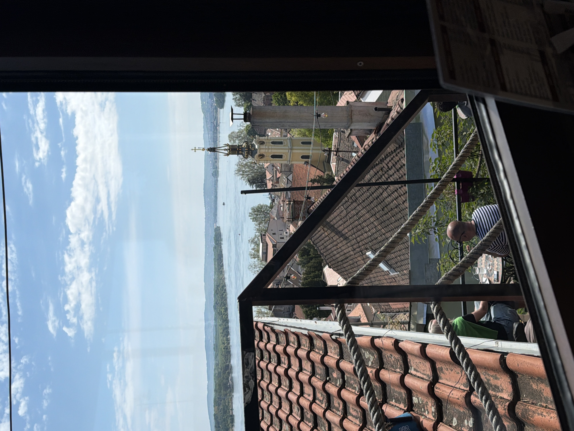 View from an elevated structure or building in Palilula Urban Municipality, Serbia, overlooking the Danube River and the rooftops of the town. A yellow church steeple with a clock is visible among the buildings. In the foreground, textured, red roof tiles and architectural elements (metal frame, rope) obscure the view of the town. Two people are partially visible; one is standing, wearing a striped shirt and looking down; another is lying down with a green garment or blanket. The sky is bright with scattered clouds.