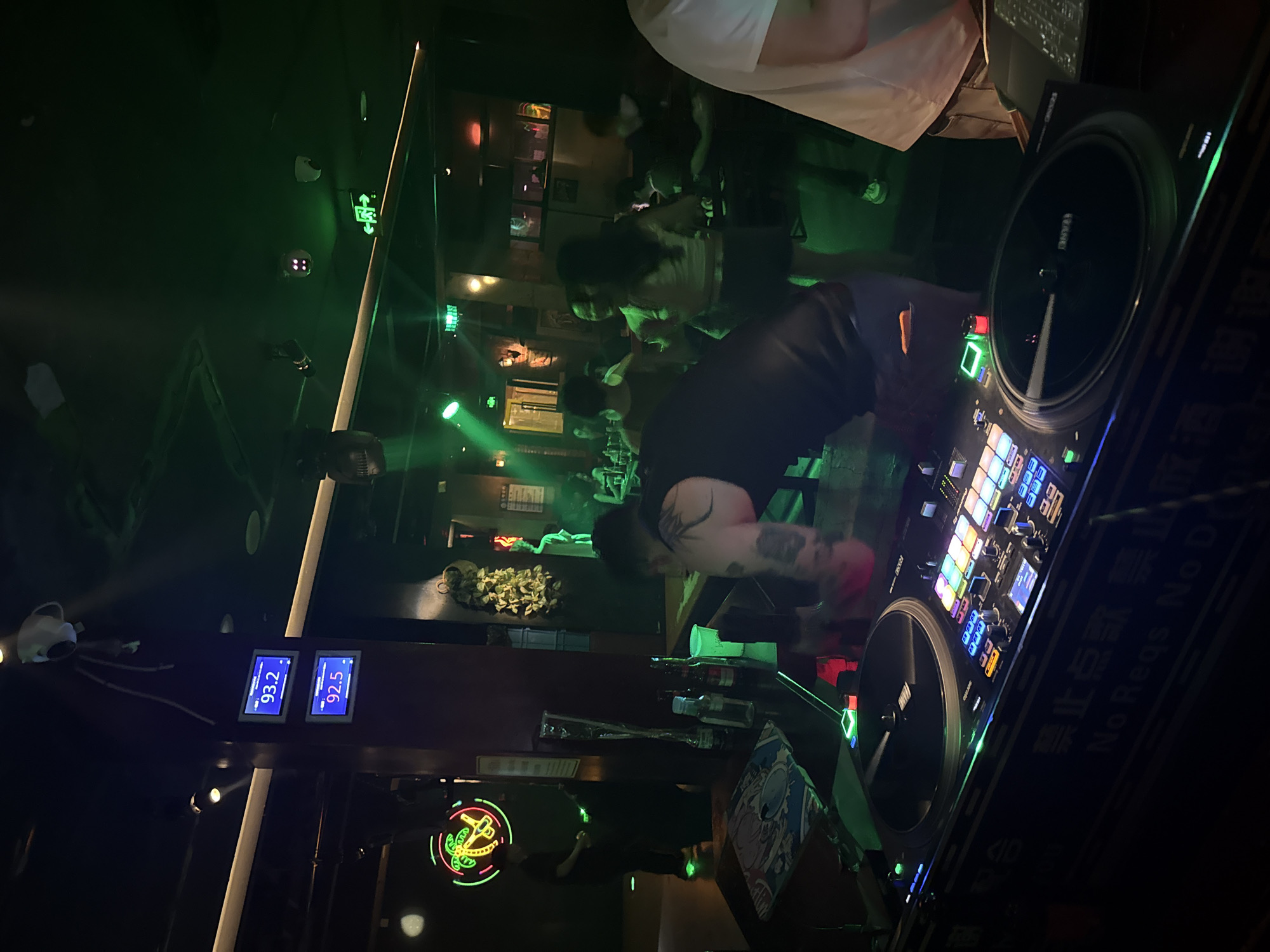 The image depicts a dimly lit nightclub or bar, likely in the city of Liangjiang, China, with green lighting dominating the scene. In the foreground, a DJ setup is visible, featuring two turntables, a mixer, and illuminated buttons. The DJ, a person with tattoos on their arm, is partially visible, appearing to be operating the equipment. In the background, several individuals are present, some appearing to be patrons enjoying themselves, with one person dancing. A neon sign depicting a palm tree is also visible on the left. The overall atmosphere suggests a lively and nocturnal social gathering.
