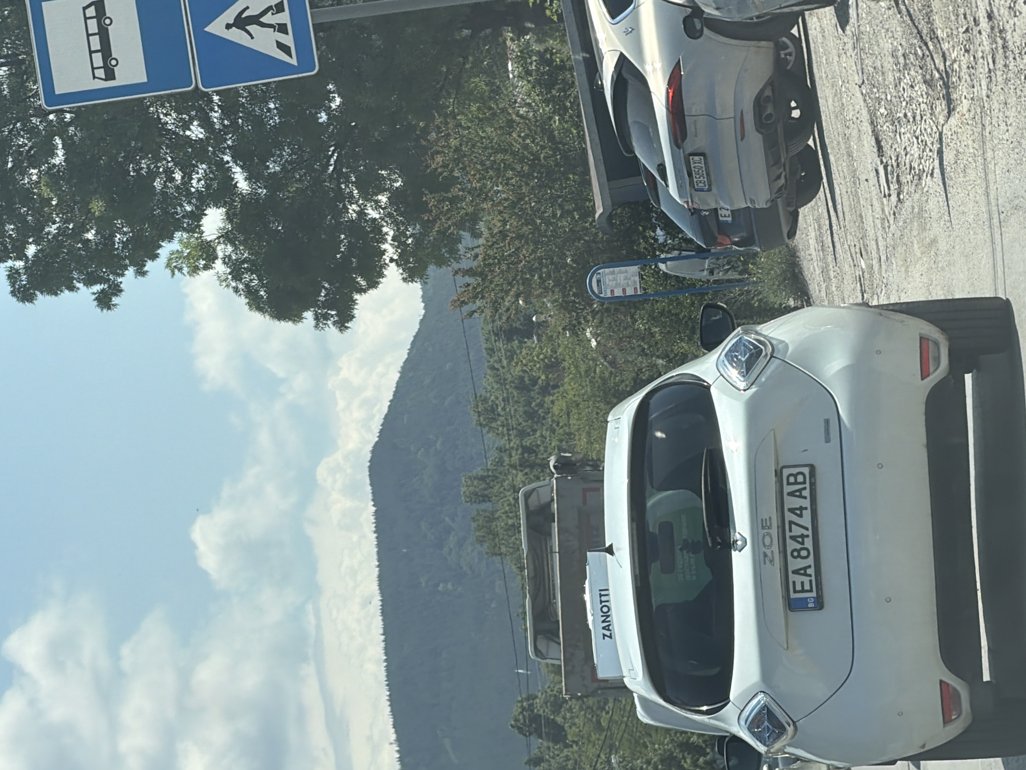 This media file depicts an outdoor scene, likely on a sunny day, with a mountain range and lush green trees in the background. The foreground features several cars, suggesting a road or parking area.
The primary subjects are the vehicles. The most prominent is a white electric car, identified by the "ZOE" badge and the license plate "EA 8474 AB". Another vehicle, a silver car, is partially visible behind it. Traffic signs are also present, including a blue sign with a white bus icon and a pedestrian crossing sign.
The setting appears to be a roadside with a gravelly surface, indicating a rural or suburban environment, consistent with the mention of Sofia, Bulgaria. The presence of trees and mountains points to a location outside the immediate urban center. The sky is bright with scattered clouds, suggesting fair weather and daylight, likely mid-morning to afternoon.
There is text visible on the vehicles and the traffic signs. The license plate of the white car reads "EA 8474 AB". The silver car has a license plate that appears to read "CB 5850 XC". A white van or truck has the brand name "ZANOTTI" printed on its side. The traffic signs are standard road signage. No people are clearly visible in the media. The overall mood is neutral, focusing on the vehicles and the environment.