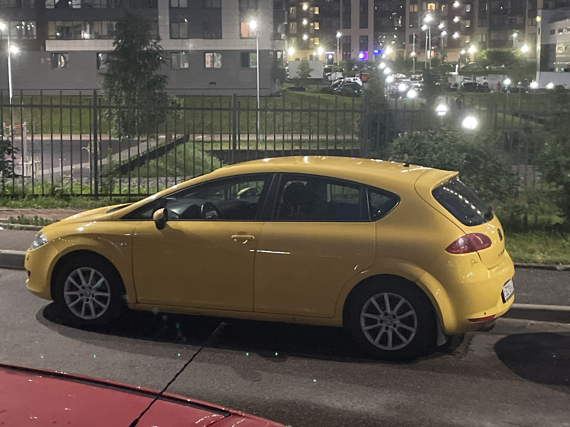 A yellow hatchback car is parked on an asphalt road at night in the city of Sinyavino, Russia. The car is viewed from the side, with its left side facing the camera. The car has five-spoke silver alloy wheels and a black tire. The car's interior is partially visible through the windows. Behind the car, a black metal fence encloses a grassy area with several trees. In the background, a multi-story apartment building is visible, with numerous illuminated windows. Streetlights are also present, casting a glow on the scene. A portion of a red car is visible in the foreground, on the lower left of the image.