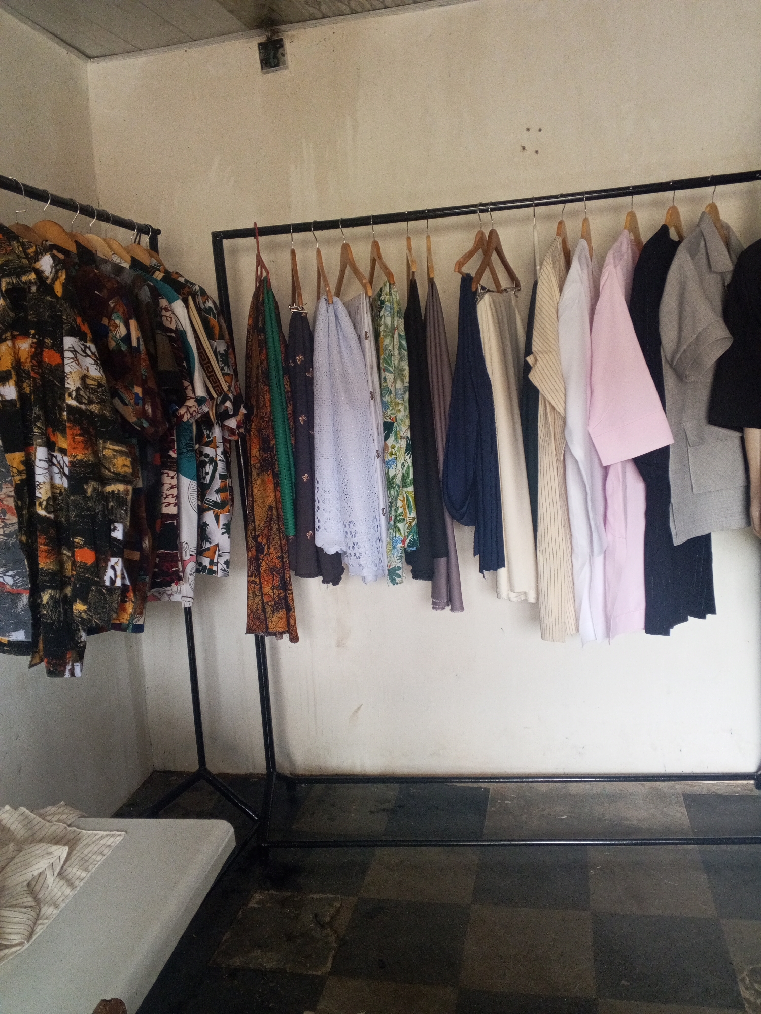 A black metal clothing rack displays an assortment of garments hanging on wooden and black plastic hangers against a textured, off-white wall. The clothes vary in pattern and color, including a brightly patterned long-sleeved shirt on the left, a white lace-trimmed garment, and several solid-colored shirts and dresses in shades of pink, grey, navy, and black. The floor beneath the rack is paved with dark and light grey checkered tiles, with some areas appearing wet or stained. To the left of the rack, a light-colored rectangular object, possibly a bed or mattress, is partially visible, with a striped fabric draped over it. The scene appears to be indoors, in a room with a simple ceiling. There are no people or animals present.