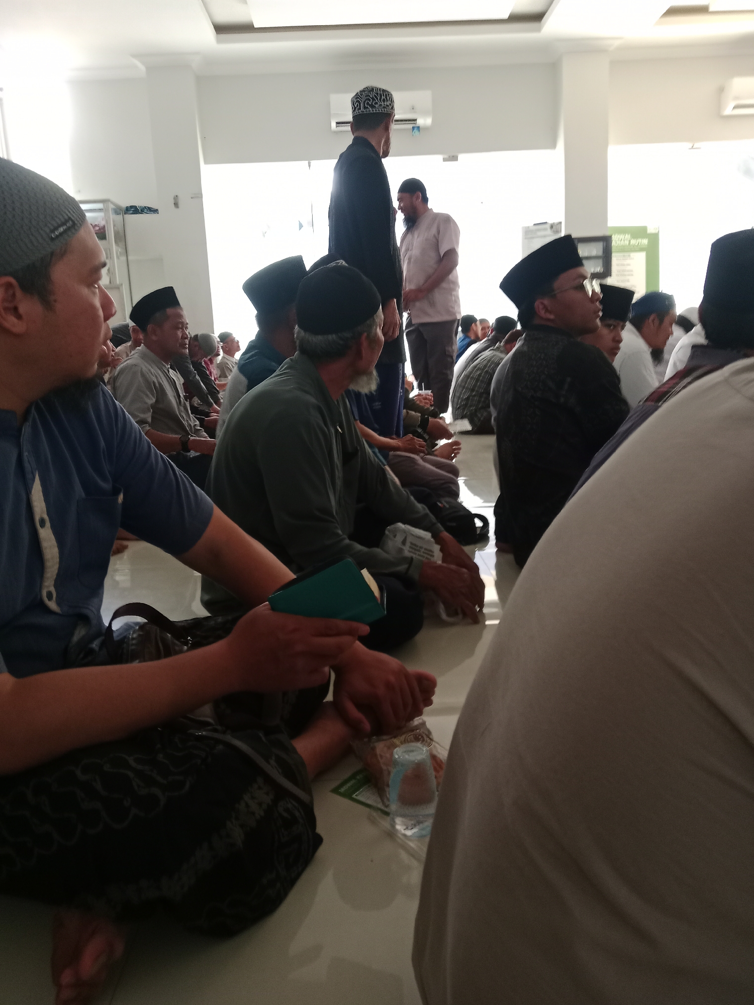 A group of men are seated on the floor of a brightly lit indoor space, likely a mosque or community hall, in Kediri City, Indonesia. The men are predominantly wearing traditional Islamic attire, including peci caps and long-sleeved shirts. Several individuals are looking in the same direction, suggesting they are listening to a speaker or observing an event. Some men are holding prayer books or small bags. The environment appears clean and well-maintained, with white walls and a polished floor. The overall atmosphere suggests a gathering for prayer or religious study.