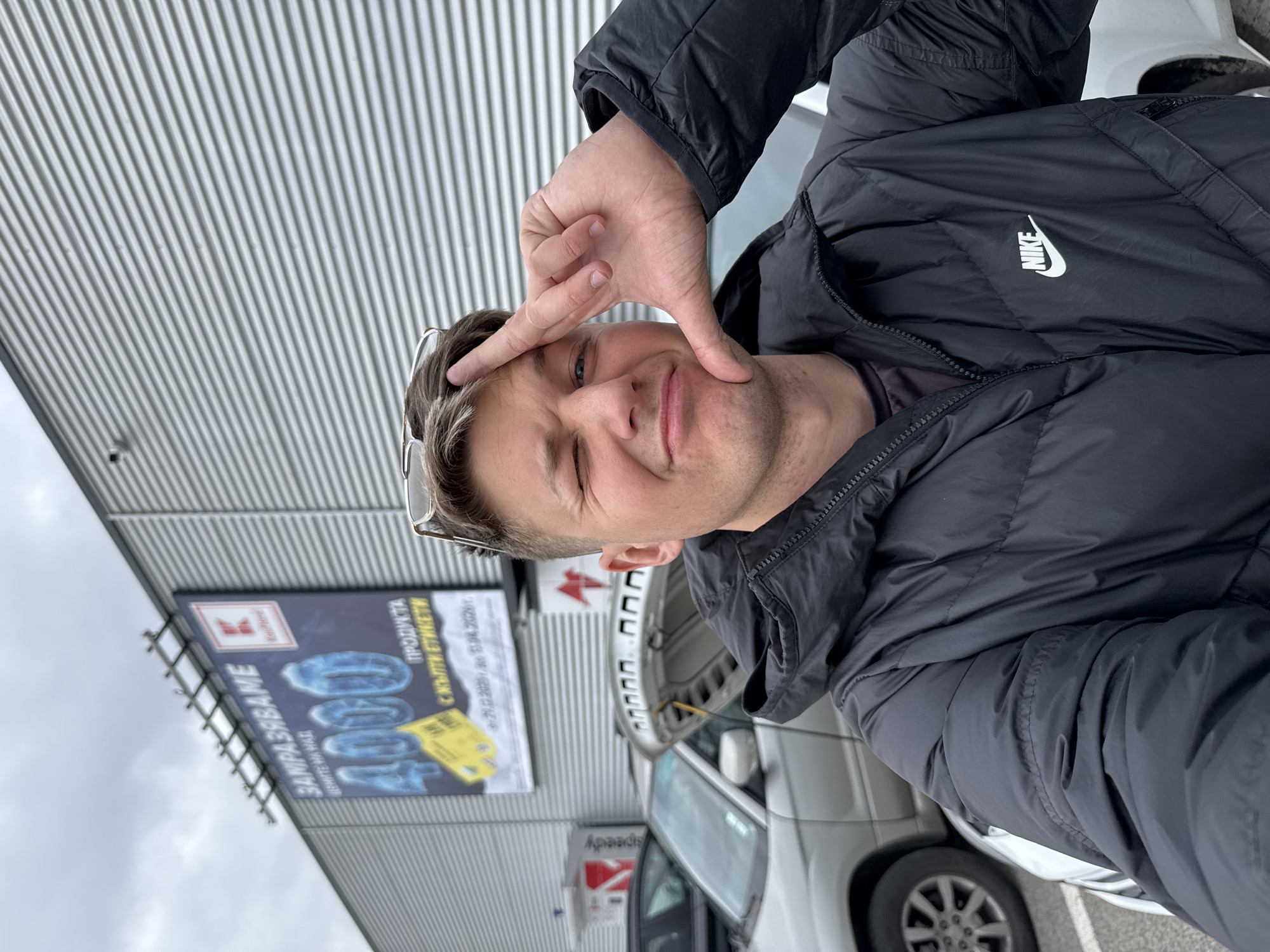 The image shows a man standing outside a building with a "Kaufland" advertisement in Burgas, Bulgaria. He is wearing a black Nike puffer jacket and glasses, and he is making a square with his hand near his face while winking. A car with its hood open is parked nearby. The location is identified by the presence of the Kaufland advertisement and the "Speedy" sign, both are common locations in Burgas, Bulgaria. The sky appears overcast.