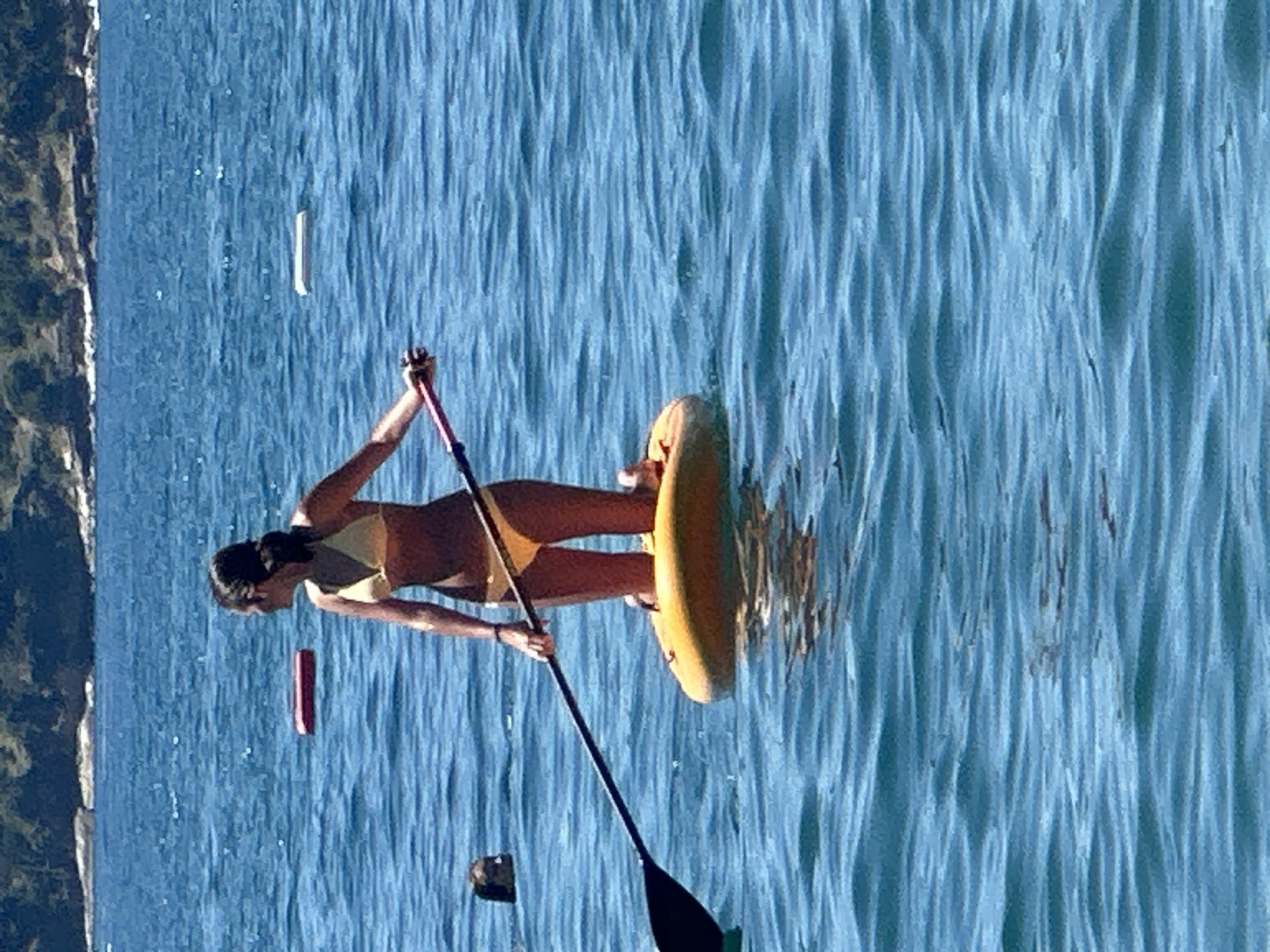 A woman is paddleboarding in the waters off the coast of Boreti, Montenegro. She is wearing a yellow bikini and is kneeling on the paddleboard. She is holding a paddle, and her arms are extended. The water is blue and calm. In the background, there is a hilly coastline covered with trees. There are other objects floating in the water, including what appear to be inflatable toys.