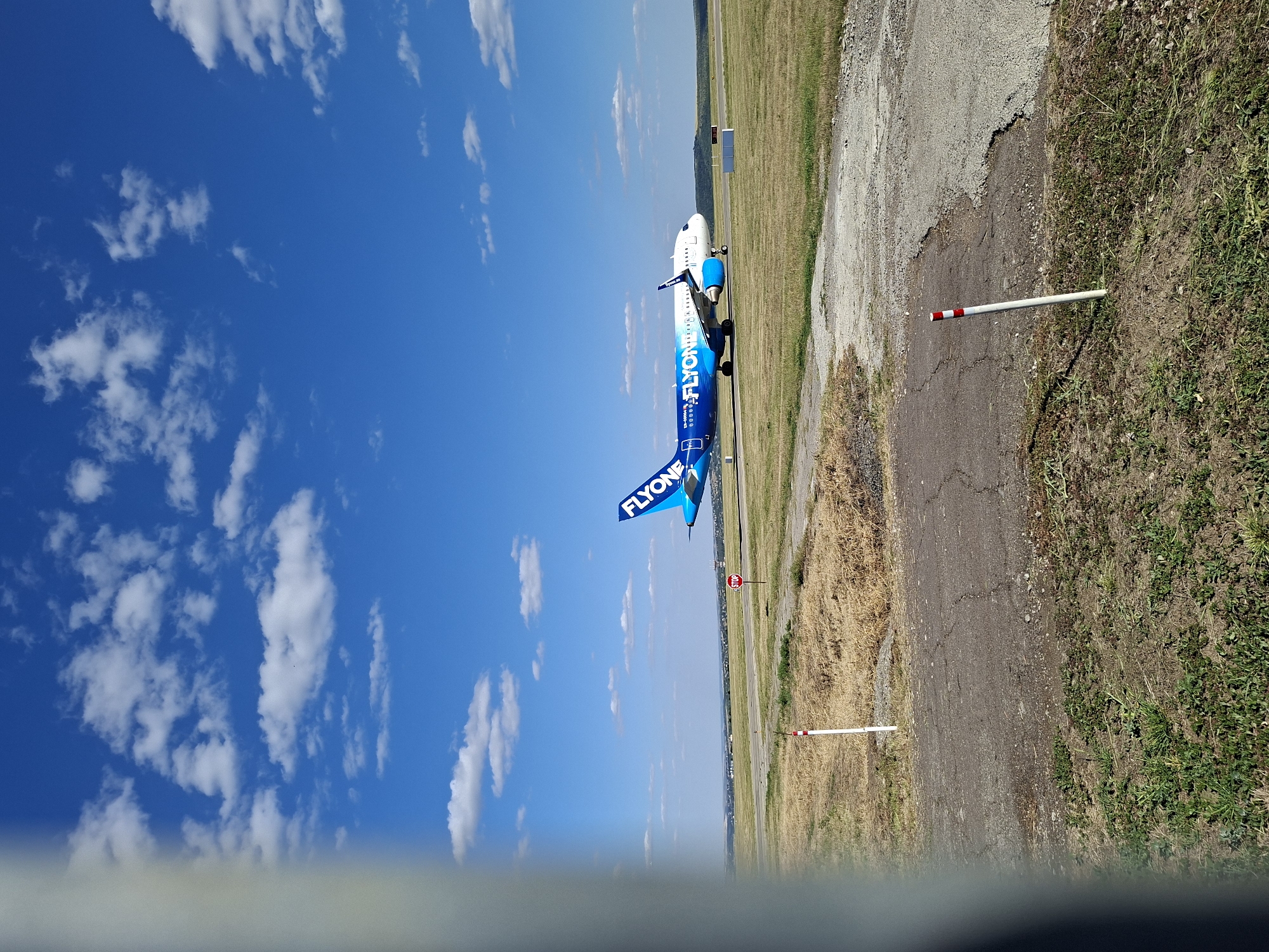 An airplane with the livery "FLYONE" on its tail is on the tarmac of Chișinău International Airport, Moldova. The airplane is blue and white, with "FLYONE" printed in white lettering on the tail. The plane is in focus, and the background is a blue sky with white clouds. The tarmac is gray, and there is dry grass on the sides. There is a red stop sign on the left side of the image. The foreground is out of focus and shows some vegetation.