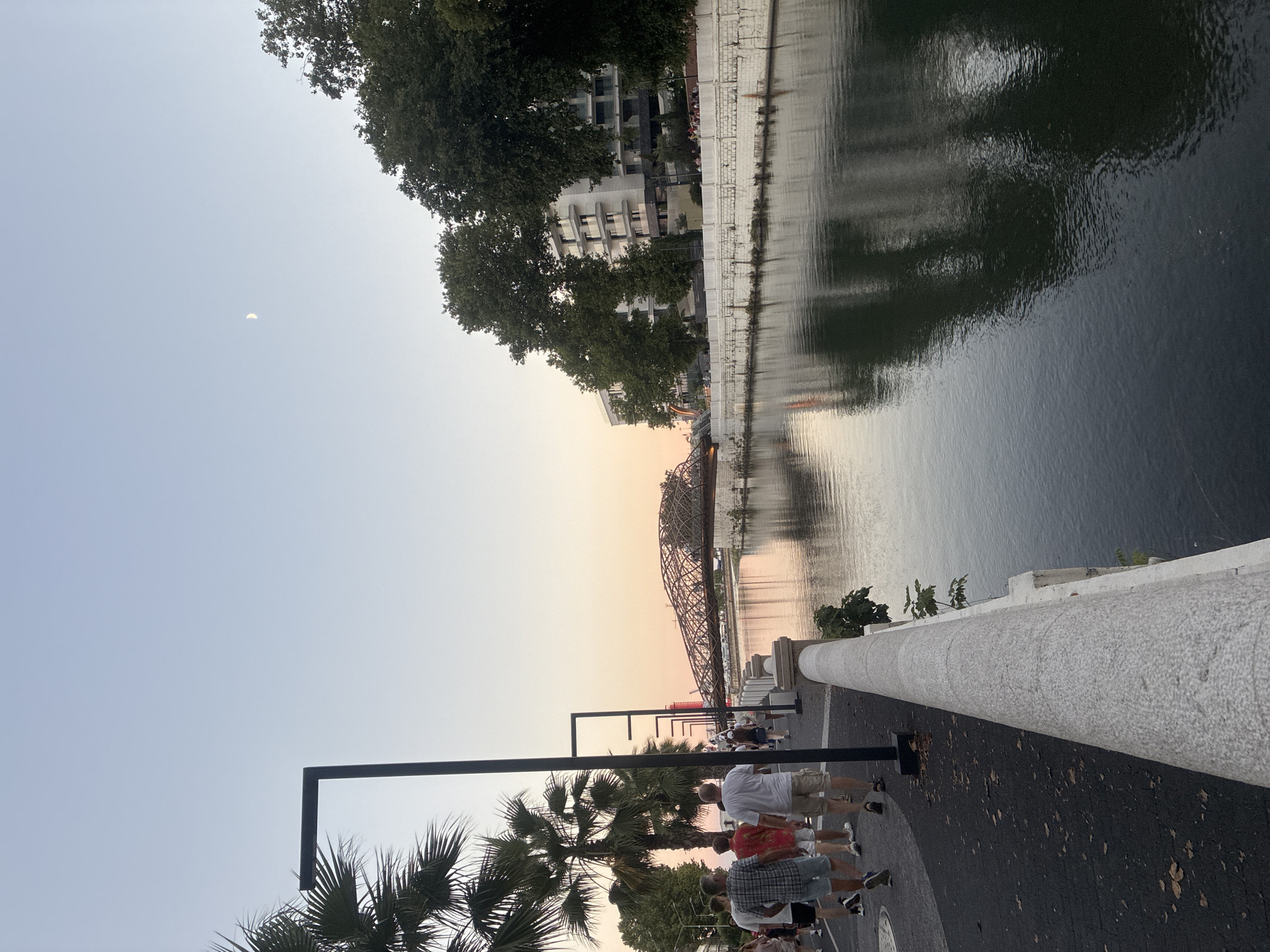 The image depicts a waterside promenade in Sochi, Russia, during dusk. A river flows alongside a paved walkway lined with palm trees and modern streetlights. Several people are walking along the promenade, some in casual attire. In the background, a distinct arched bridge spans the river, and modern buildings with balconies are visible on the opposite bank. The sky is a pale gradient of blue and pink, with a sliver of the moon visible. The water reflects the ambient light and the surrounding architecture.