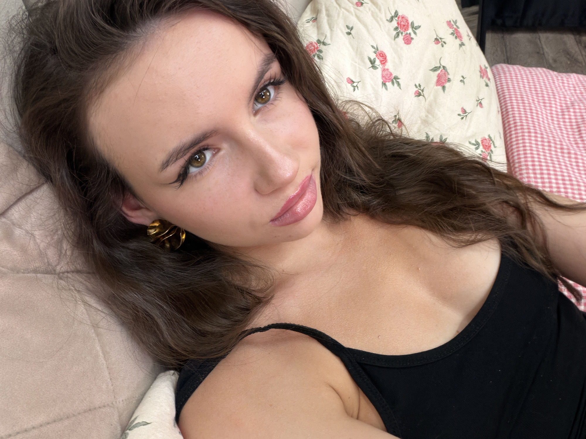 This is a close-up, selfie-style photograph of a young woman lying down. She has long, dark brown hair, light brown eyes, and is wearing makeup, including winged eyeliner and lip gloss. She is wearing a black tank top. Her right ear is adorned with a large, gold, textured earring. The background suggests a casual, indoor setting, possibly a bedroom or living room, with bedding that includes a floral pattern and a pink gingham print. The lighting appears to be soft and natural, suggesting it could be daytime. The woman's expression is relaxed and somewhat playful, with a slight smile. There is no visible text in the image. The location context provided is Chaikovsky, Russia, but this cannot be confirmed from the image alone.